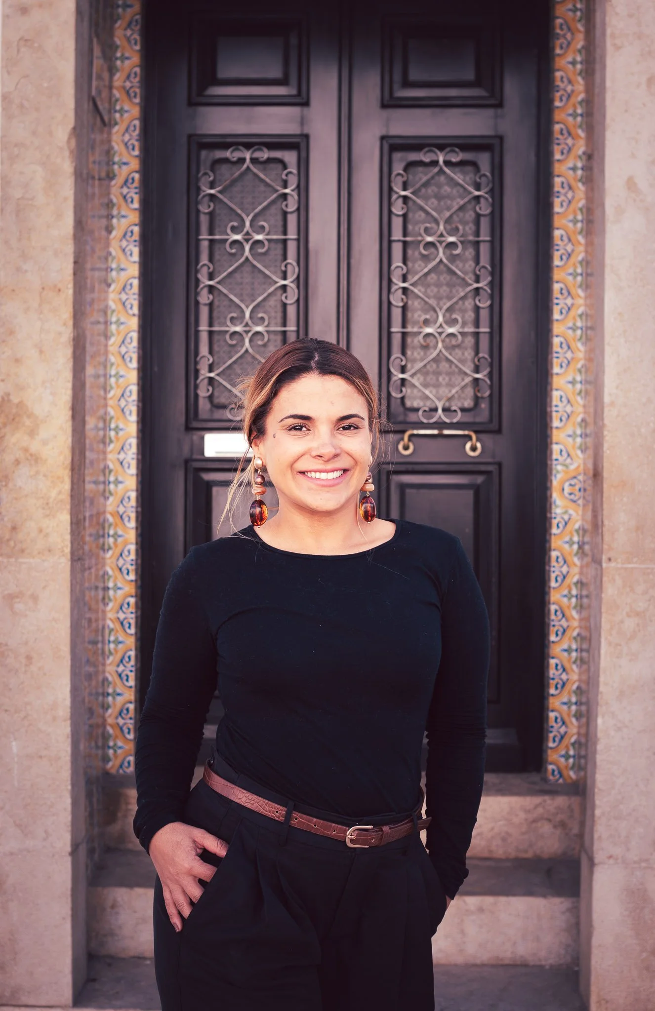 A woman with brown hair, wearing a black long-sleeve shirt and black pants with a pink belt, smiling and standing in front of a dark wooden door with decorative ironwork and tile accents.