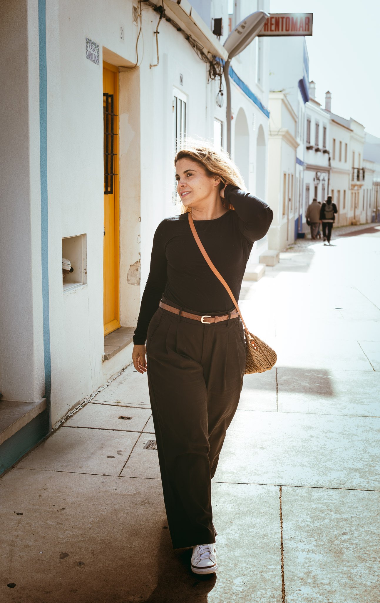 A woman walking on a sunny street with pastel-colored buildings, wearing a black long-sleeve shirt, dark pants, white sneakers, and carrying a tan crossbody bag.