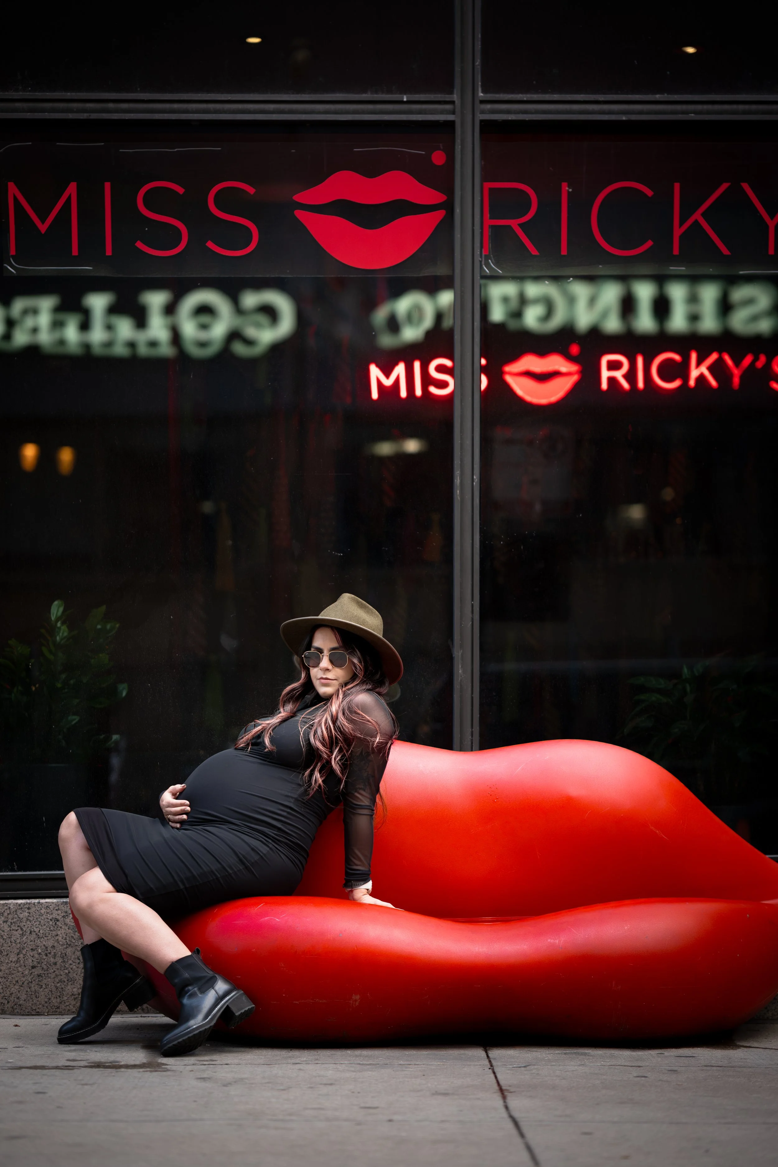 A pregnant woman with long hair, sunglasses, black dress, black boots, and a hat sitting on a large red lips-shaped bench outside a storefront with neon signs reading Miss Ricky's.