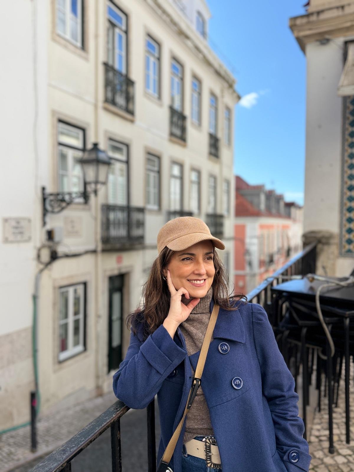 Smiling woman in a blue coat and beige hat standing on a balcony with a city street behind her.