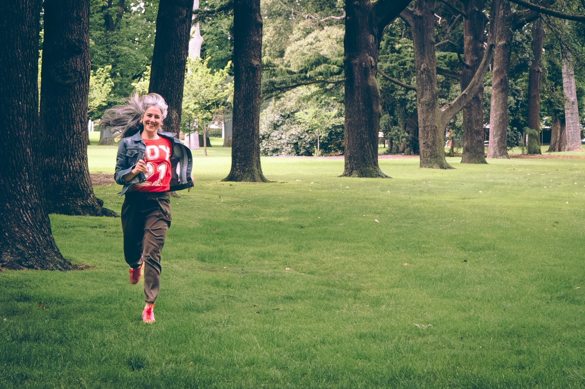 Woman with gray hair running happily through a park with green grass and tall trees.