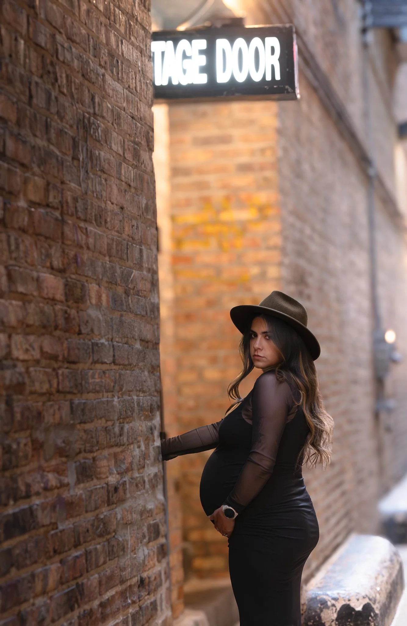 A pregnant woman with long wavy hair wearing a black dress and a brown wide-brimmed hat standing against a brick wall on a city street, looking at the camera with a serious expression. There is a lit sign that reads 'VINTAGE DOOR' above her.