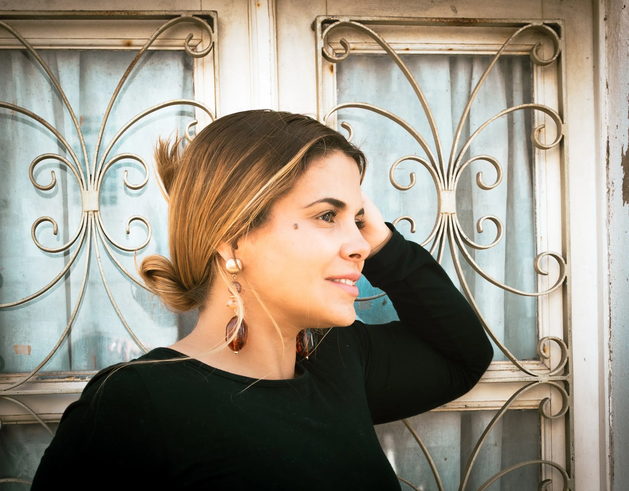 A woman with long hair tied in a bun, wearing earrings and a black top, standing in front of an ornate metal window grille, turned sideways with a gentle smile.