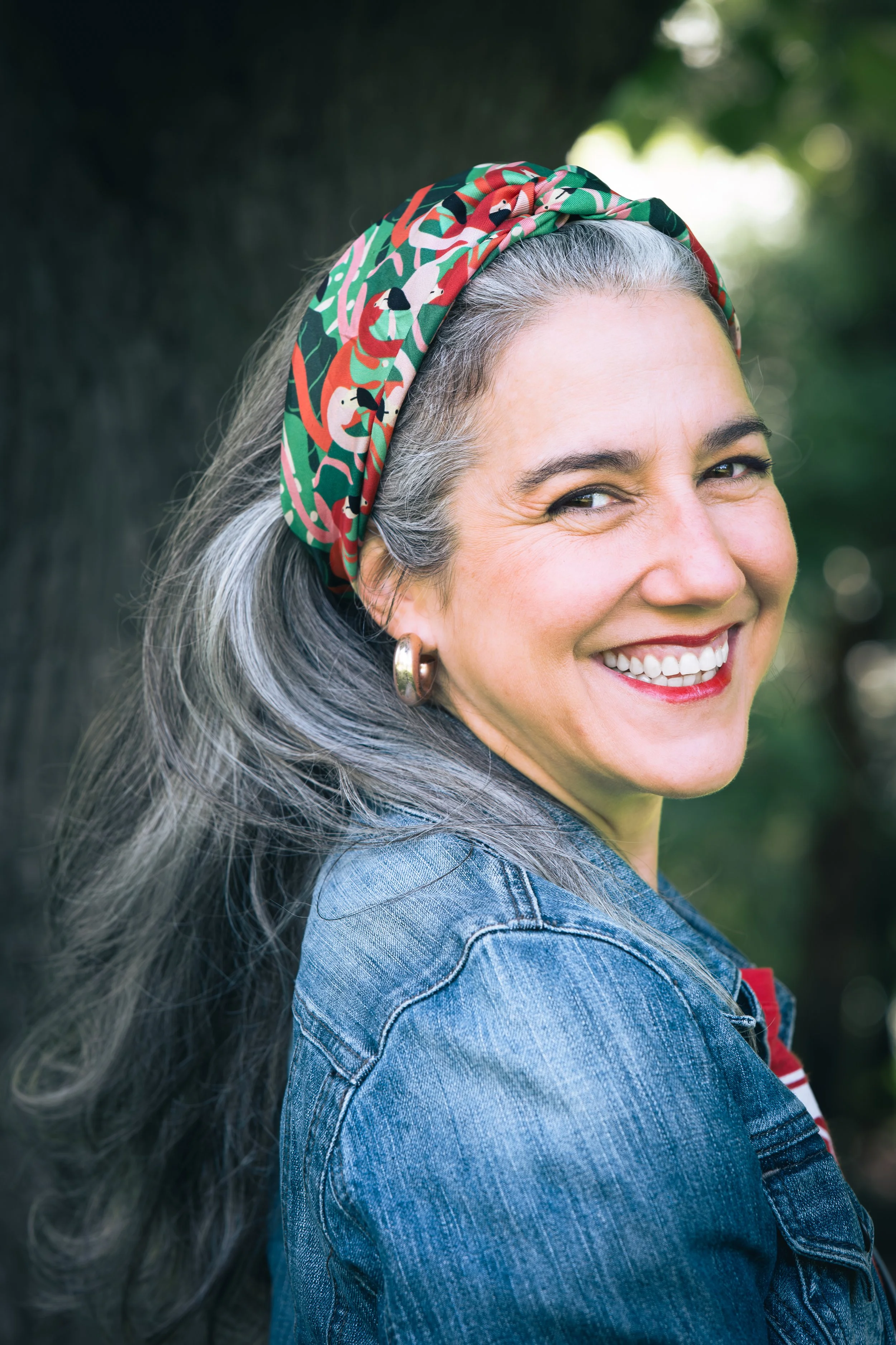 Smiling woman with long gray hair wearing a colorful headscarf, hoop earrings, and a denim jacket outdoors.