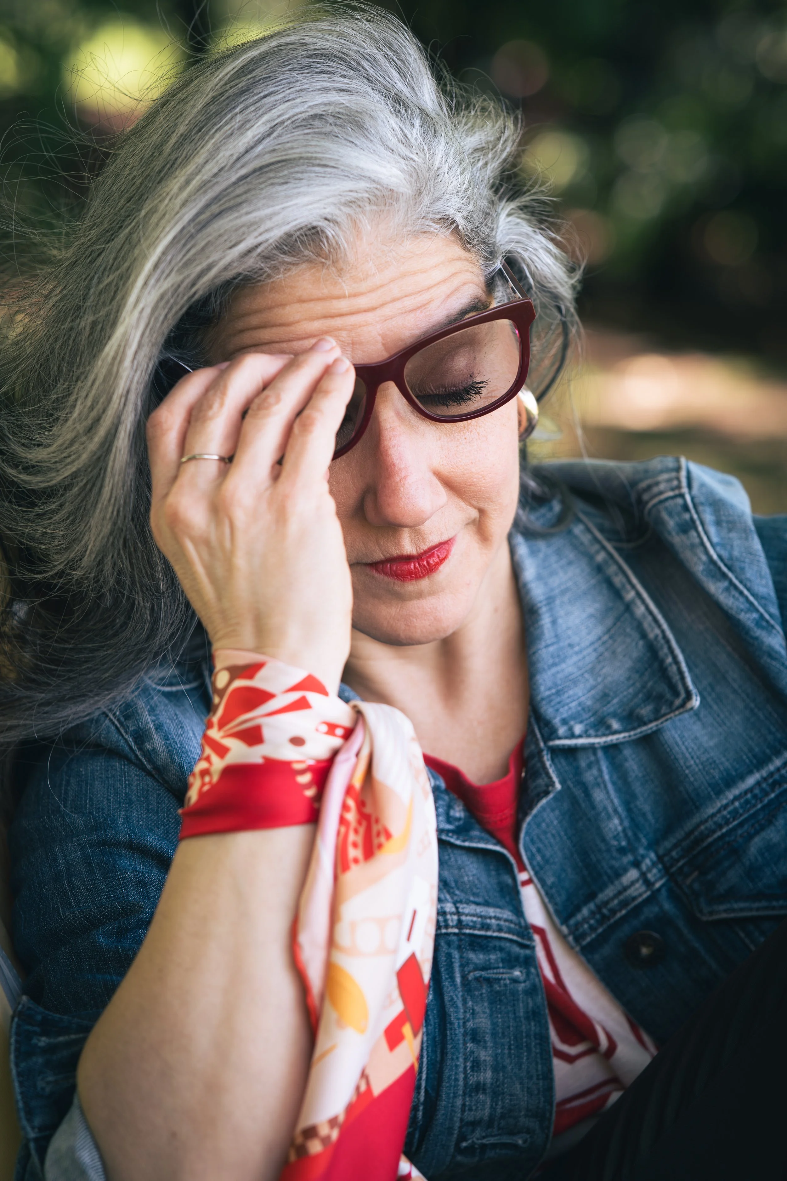 A woman with gray hair and red glasses sitting outdoors, touching her forehead, with a pensive expression, wearing a denim jacket and a colorful scarf.