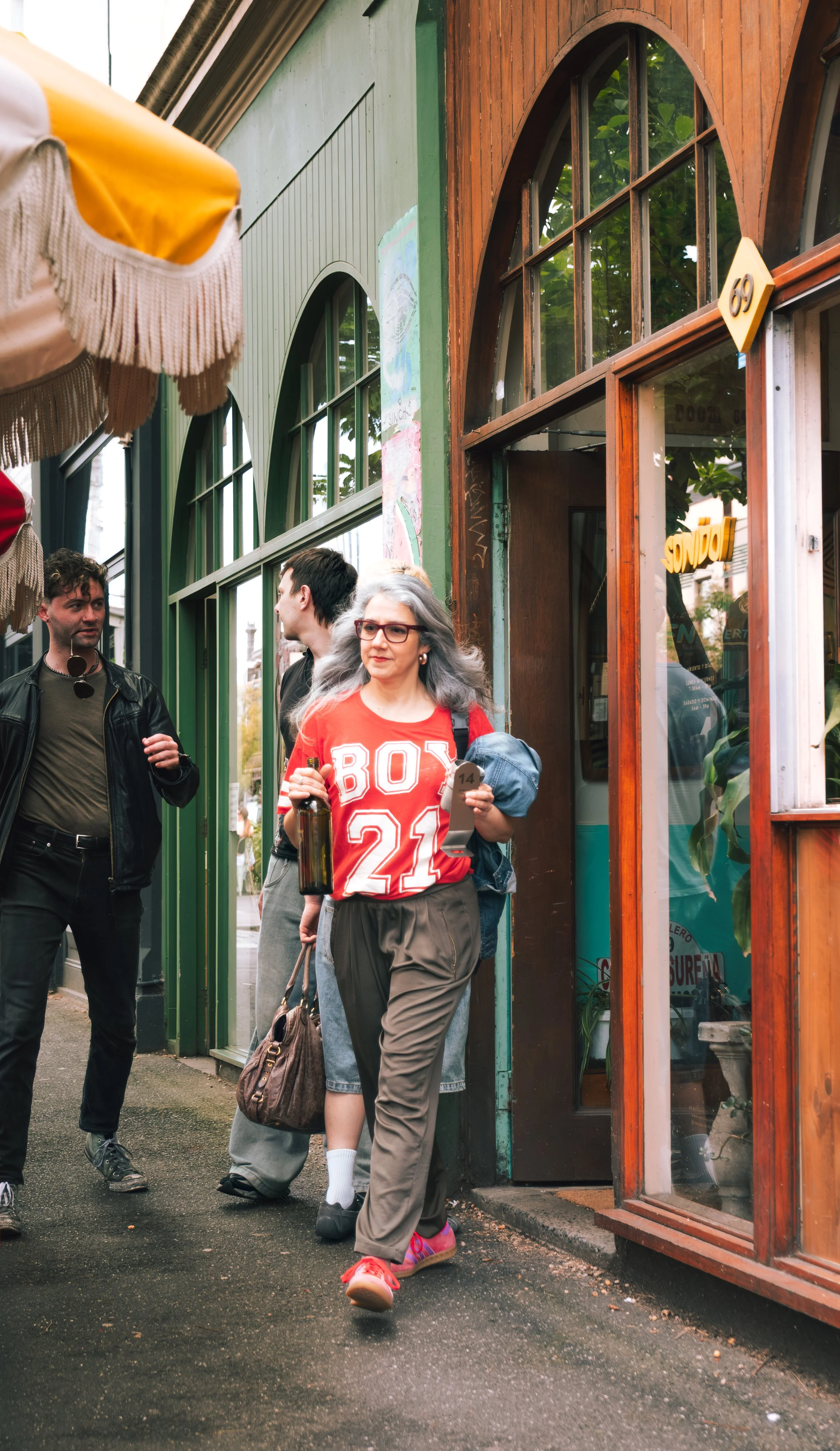 People walking past a storefront with green and wood trim, chatting, while one woman in a red shirt with 'BOY 21' on it carries a bottle and a towel.