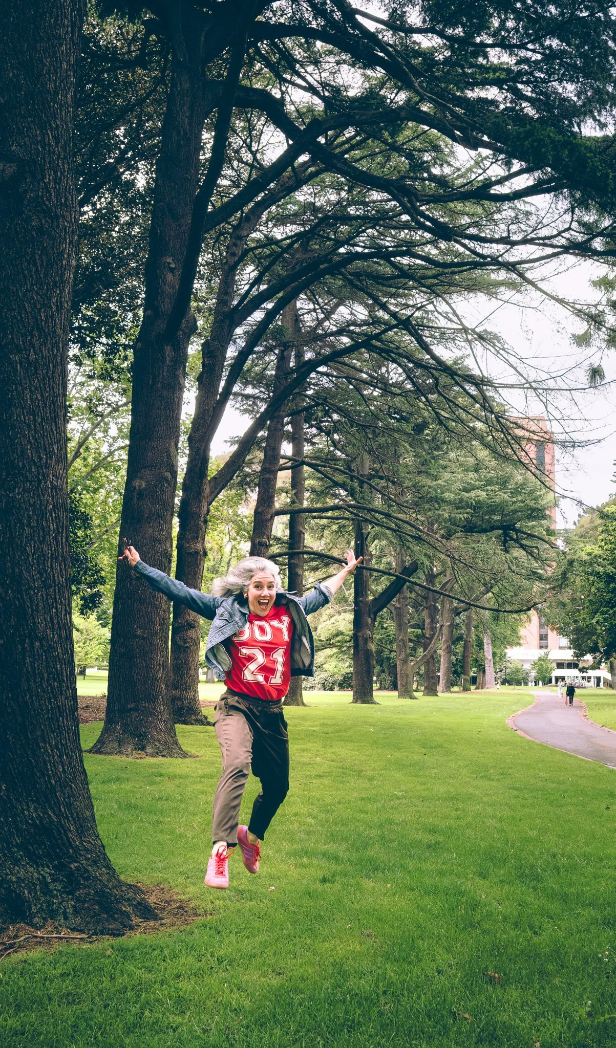 A woman jumping joyfully next to large trees in a park with a curved walking path and tall buildings in the background.
