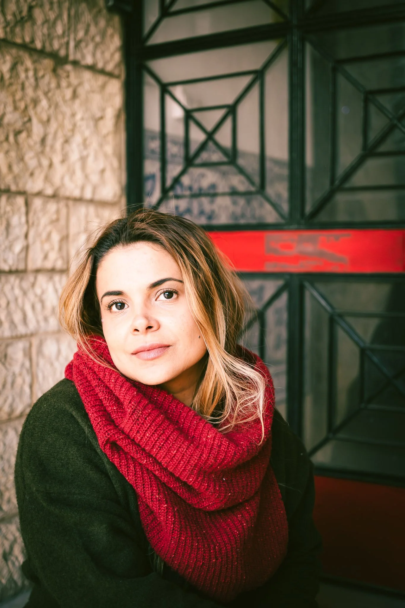A woman with light brown hair and fair skin wearing a red knit scarf and a dark coat, sitting in front of a stone wall and glass door with black frame.