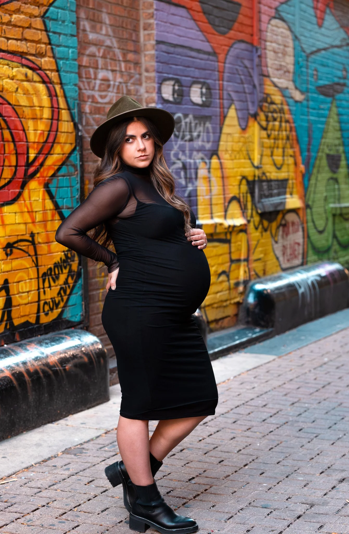 A pregnant woman in black dress and black boots standing against a colorful graffiti wall on a brick sidewalk.