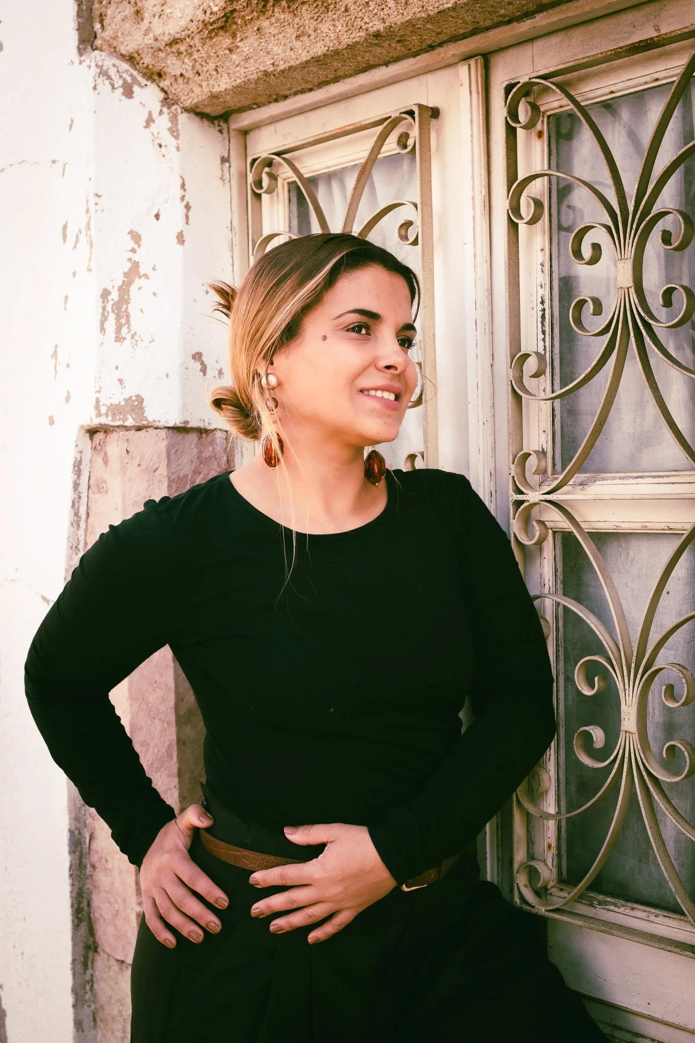 A woman with light brown hair tied back, wearing a black long-sleeve shirt and large earrings, stands outdoors near a decorative wrought iron window grill, smiling and looking to her right.