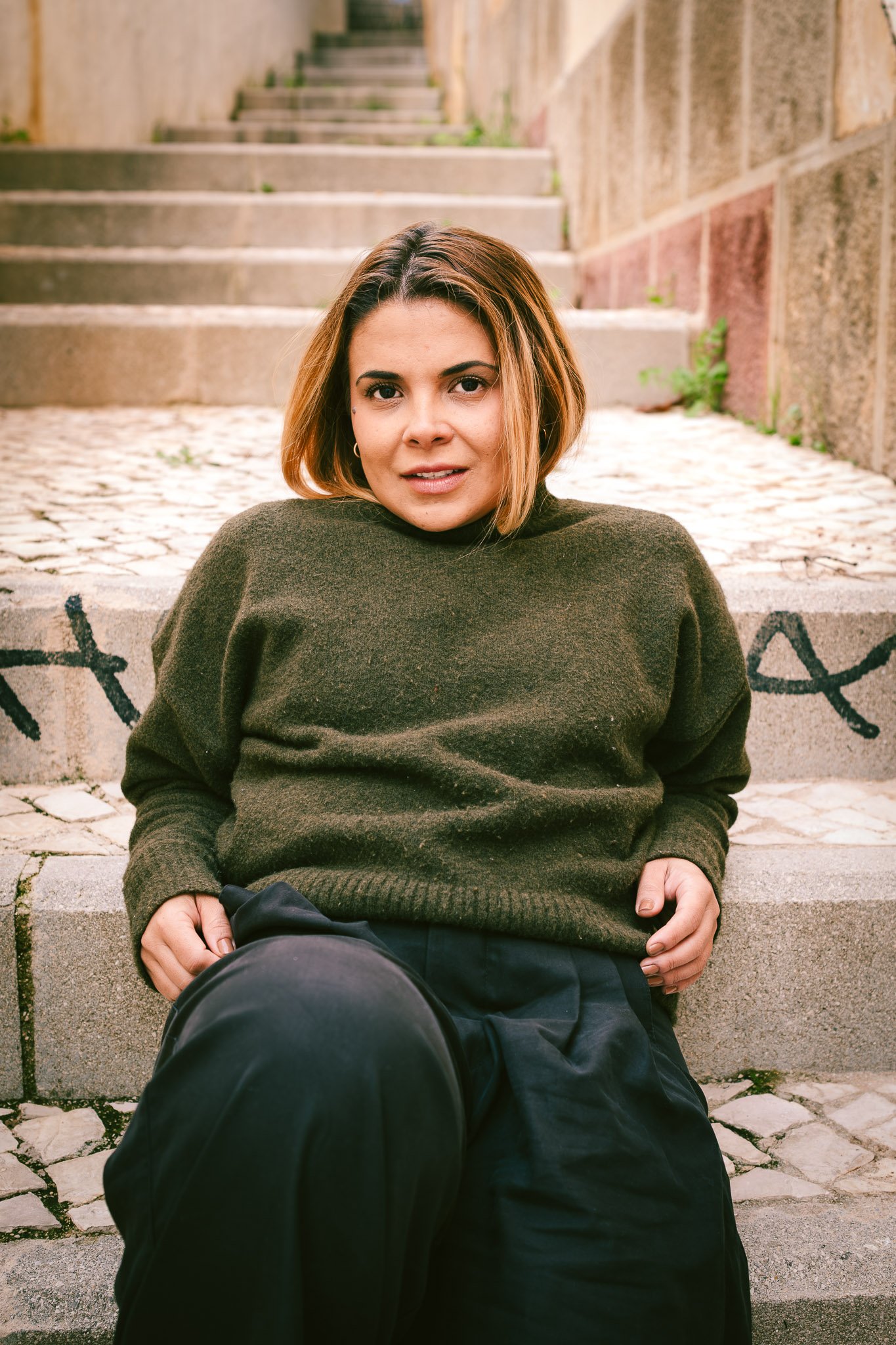 A woman with short, light brown hair sitting on outdoor stone steps, looking at the camera. She is wearing a dark green sweater and black pants, with graffiti on the step behind her.