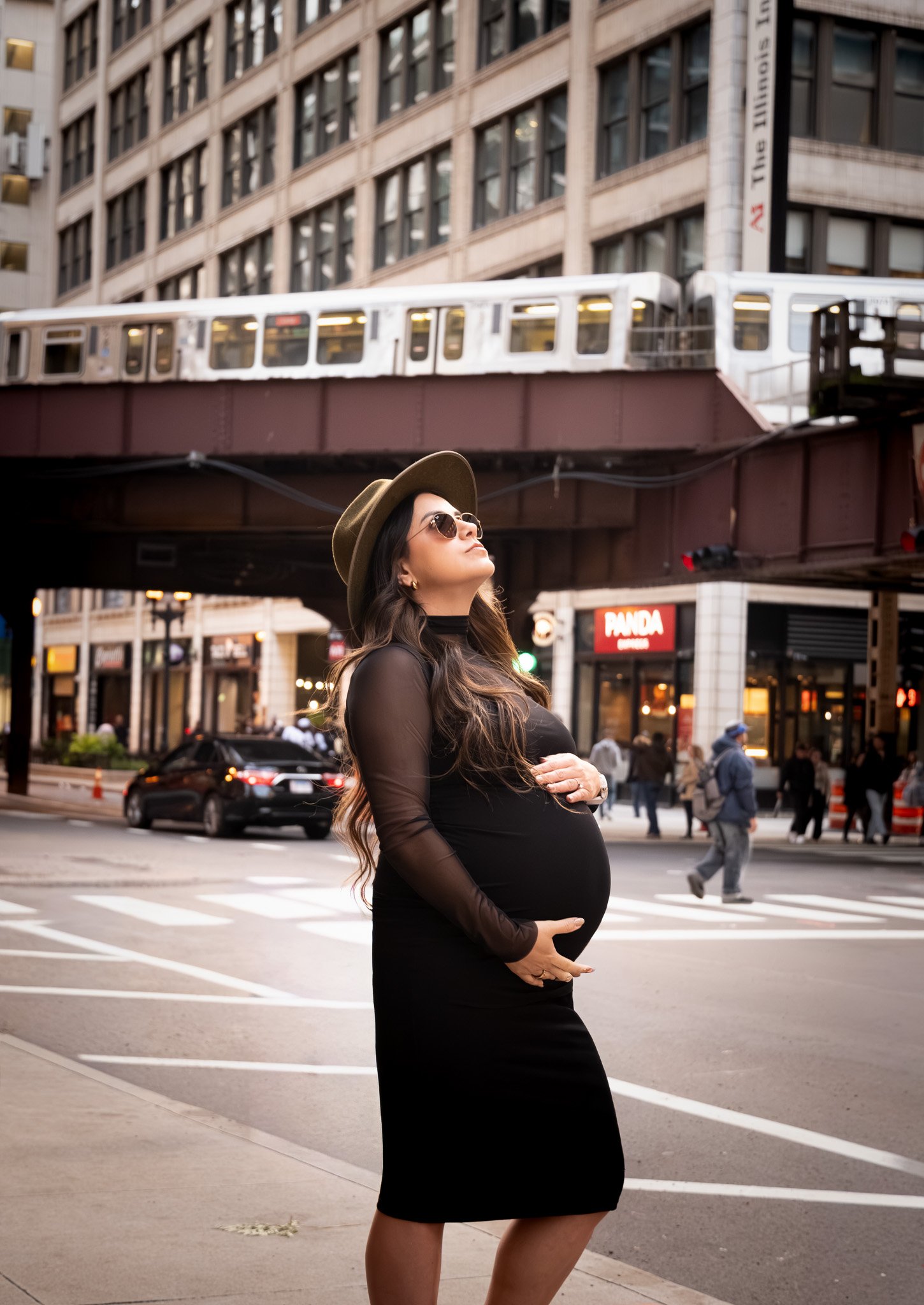 Pregnant woman standing on city street corner wearing sunglasses, hat, black dress with sheer sleeves, holding her belly, with city buildings, cars, and pedestrians in the background.