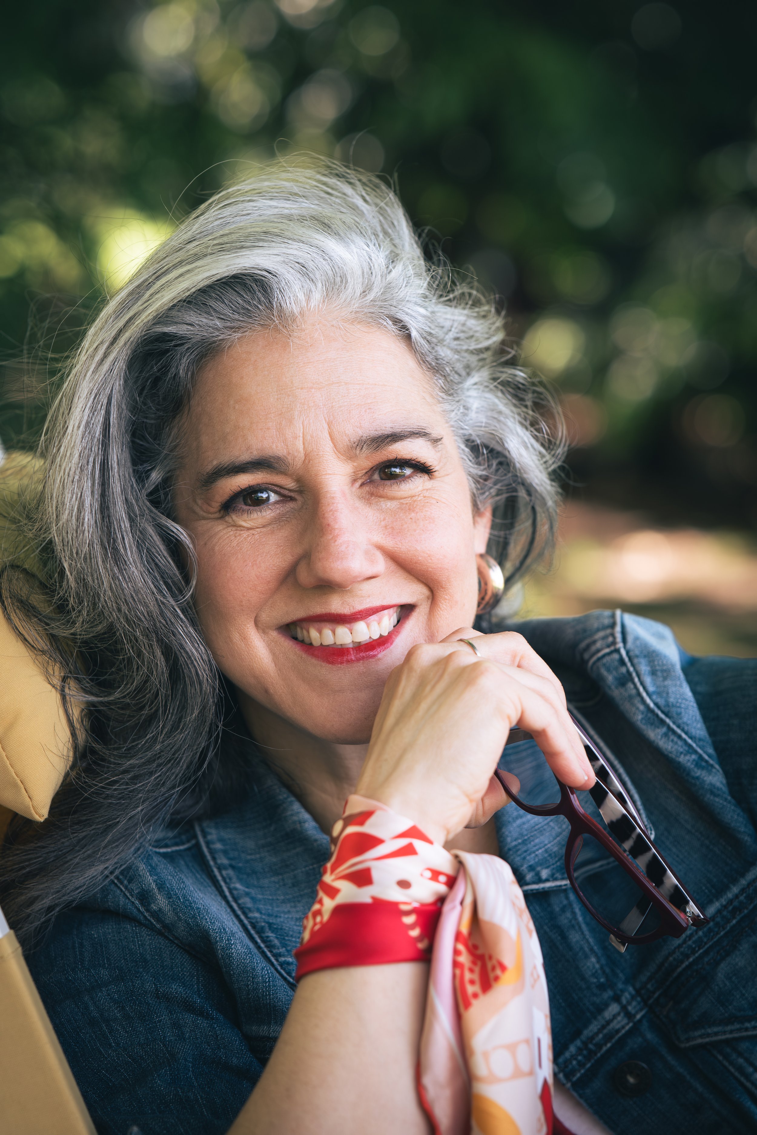 A smiling woman with gray hair and freckles, wearing a denim jacket, red lipstick, and a colorful scarf, outdoors with blurred green foliage in the background.