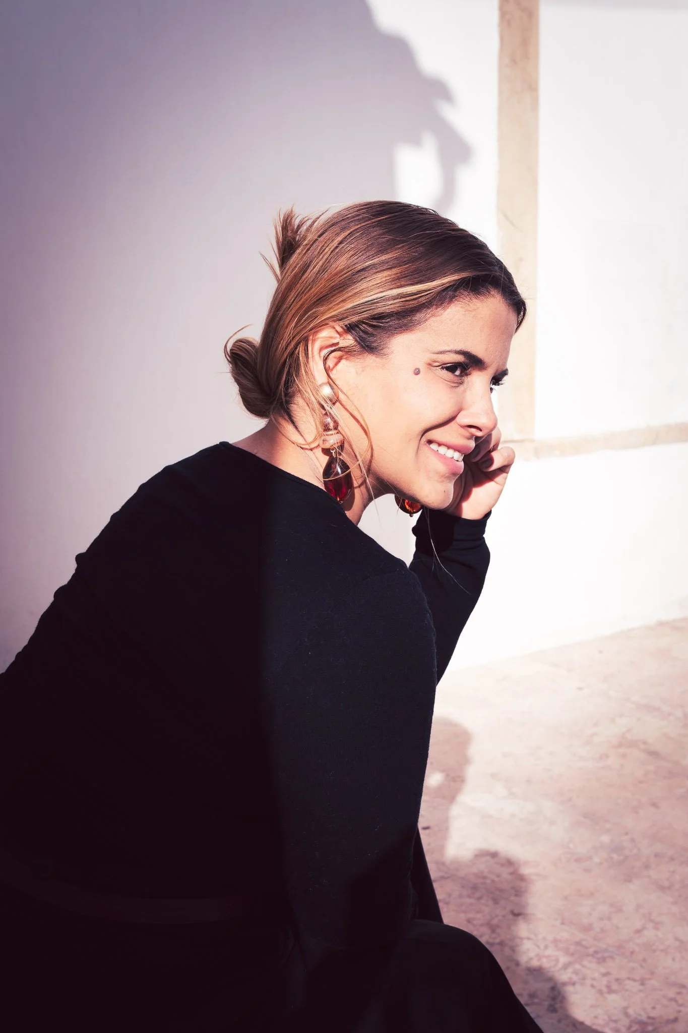 A woman with short, light brown hair talking on a cell phone, sitting next to a white wall with a shadow cast on it, smiling and wearing earrings and a black long-sleeve shirt.