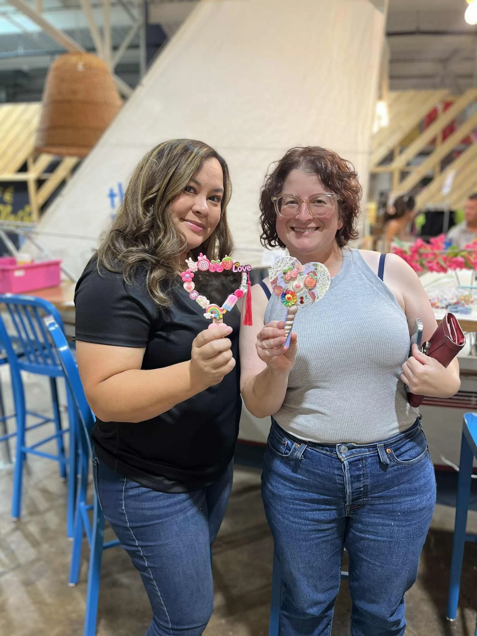 Two women smiling and holding decorated heart-shaped cookies at a bakery or craft fair, with colorful chairs and display tables in the background.