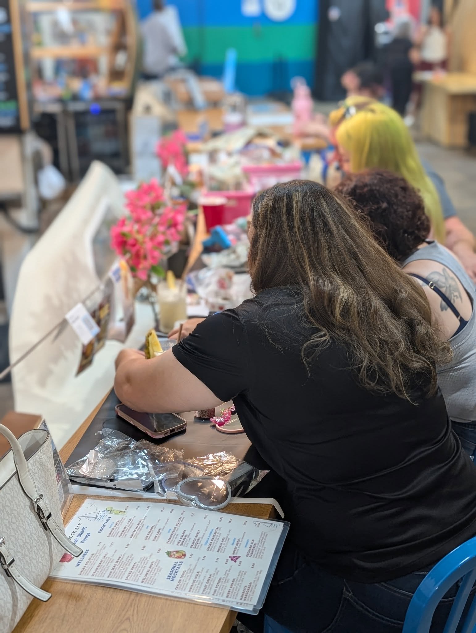 A group of people sitting at a long table in a busy indoor market or food hall, with one woman in a black shirt in the foreground, reading a menu, and others engaging in conversation or looking at items on the table.