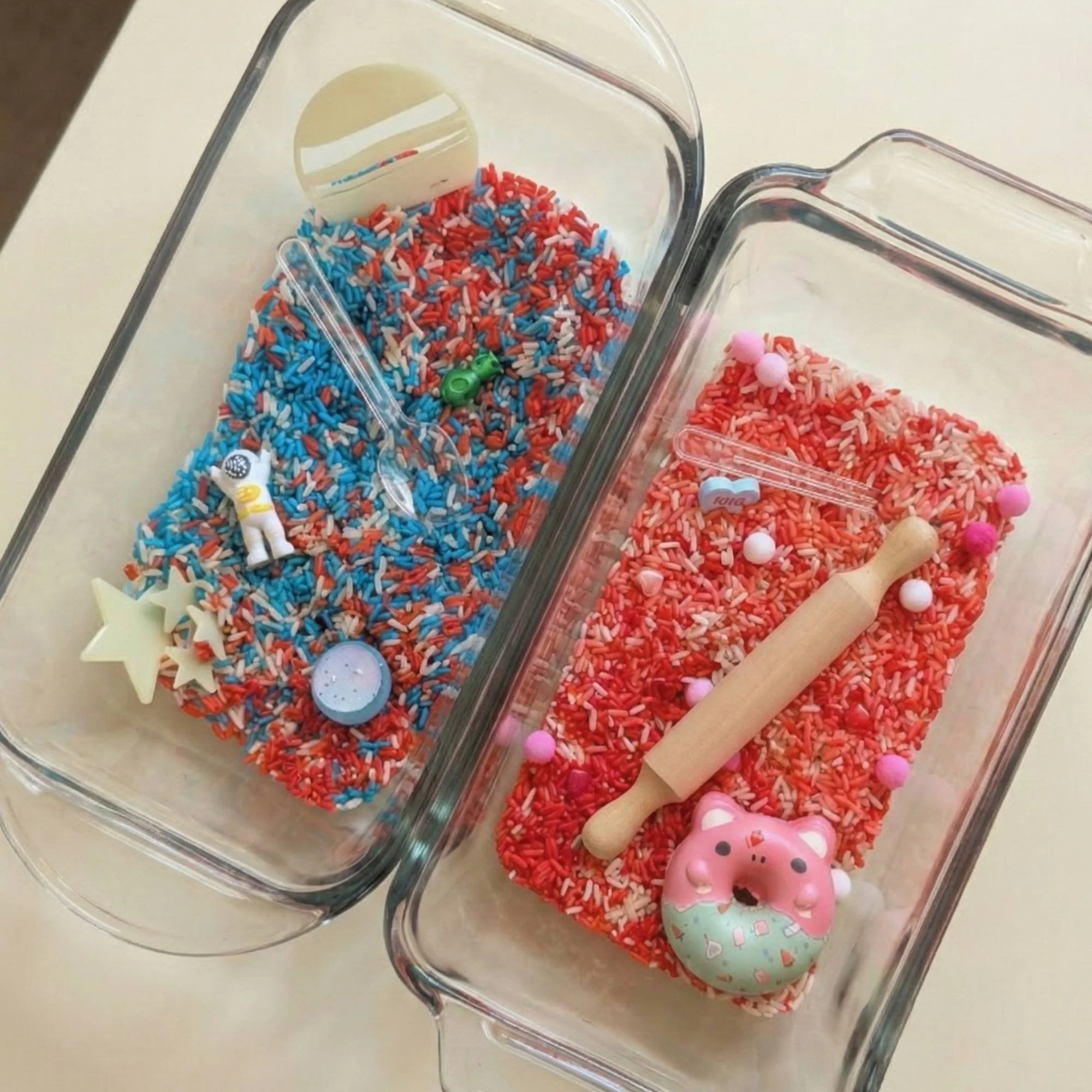 Two glass containers filled with red, white, and blue sprinkles. The left container has small space-themed toys like a miniature astronaut, stars, a rocket, and a round light. The right container has a pink pig-shaped figurine, a small rolling pin, and pink pom-poms along with the sprinkles.