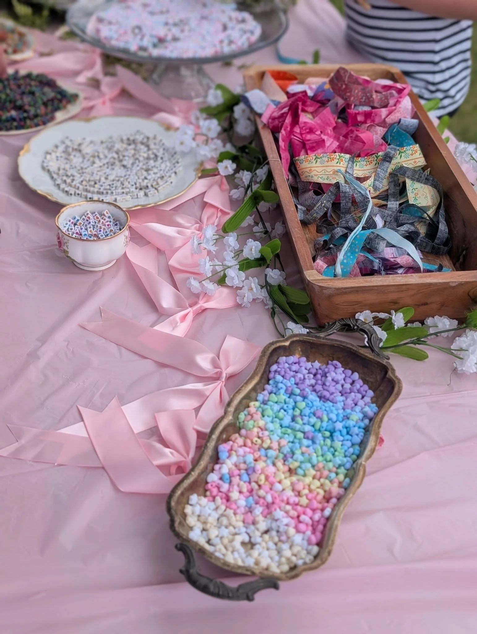 A table decorated with pink tablecloth, pink ribbons, and white flowers, displaying colorful beads, ribbons, and small craft supplies for a craft or jewelry-making activity.