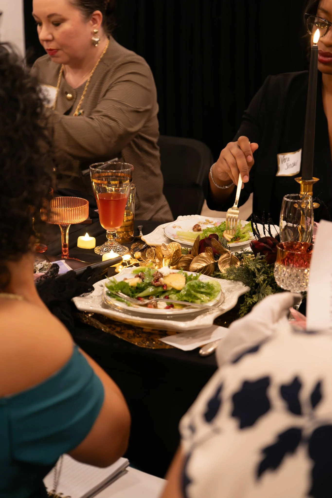A dinner party with women seated around a table adorned with plates of salad, wine glasses, candles, and holiday decorations.