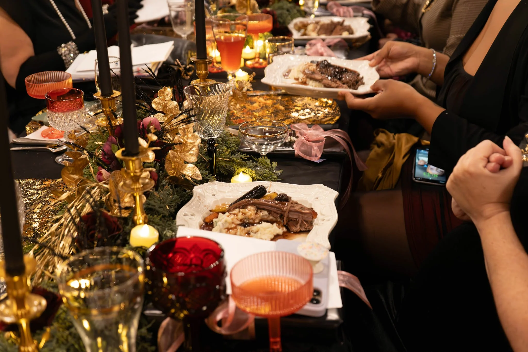 A holiday dinner table decorated with candles, a garland centerpiece, wine glasses, and plates of food, with people serving themselves and sitting around the table.