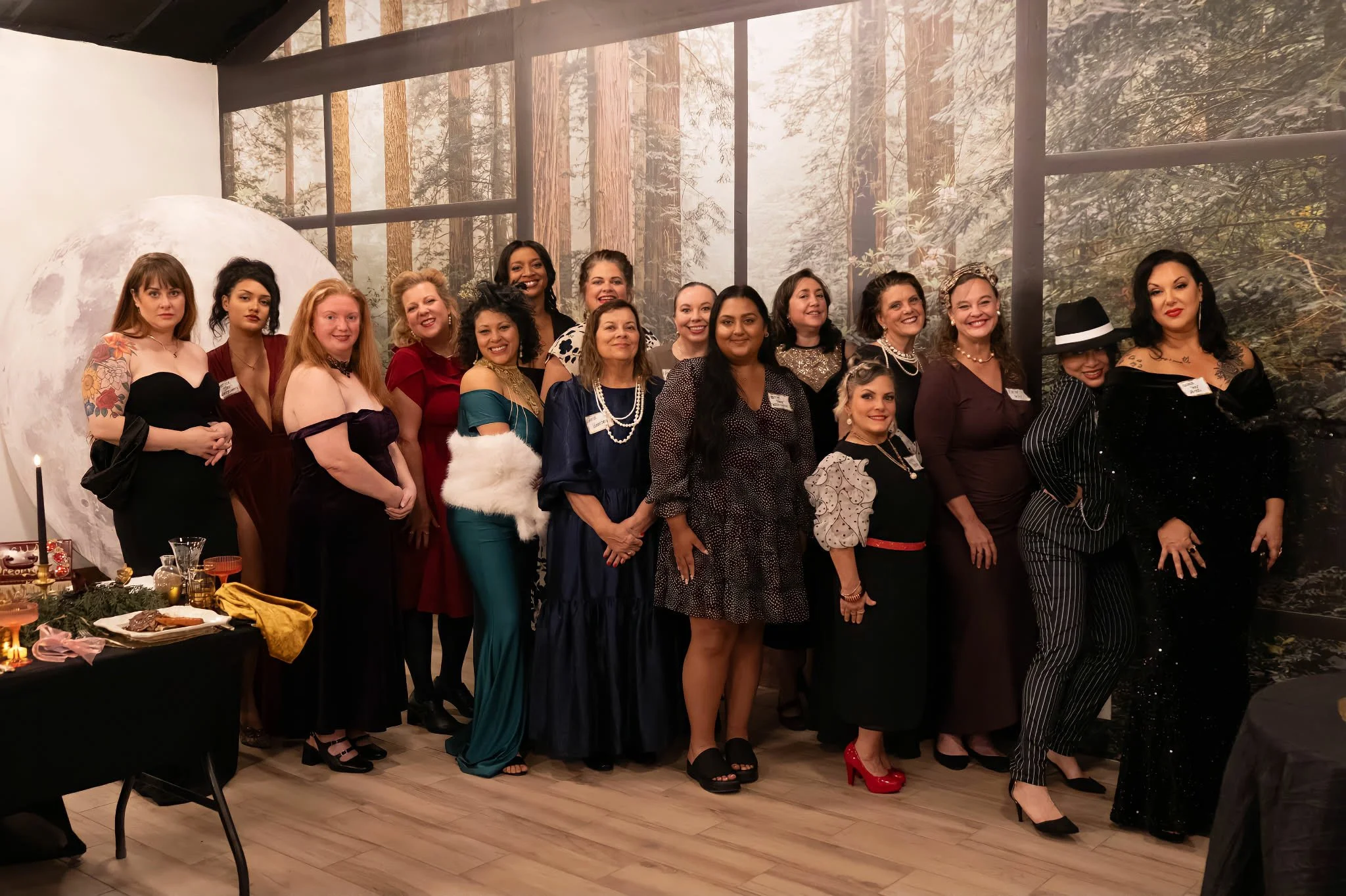 Group of women dressed elegantly at a party in a room with forest-themed wall decor, with a moon scene and a table with candles and food on the side.