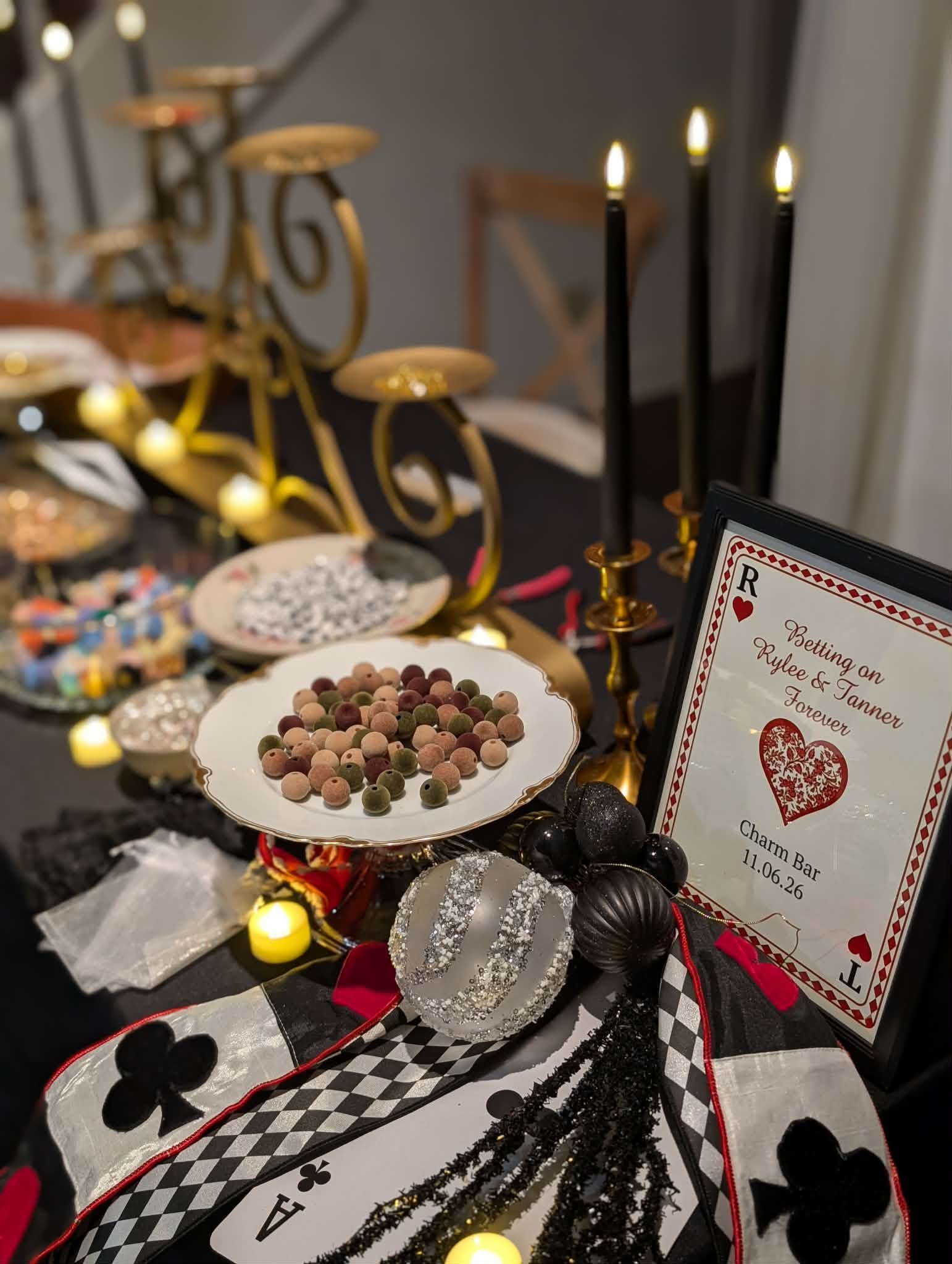 Table with casino-themed decorations including black and red playing cards, a framed picture reading 'Betting on Rylee & Tanner Forever,' candles, candies, and black ornaments, set for a celebration or party.