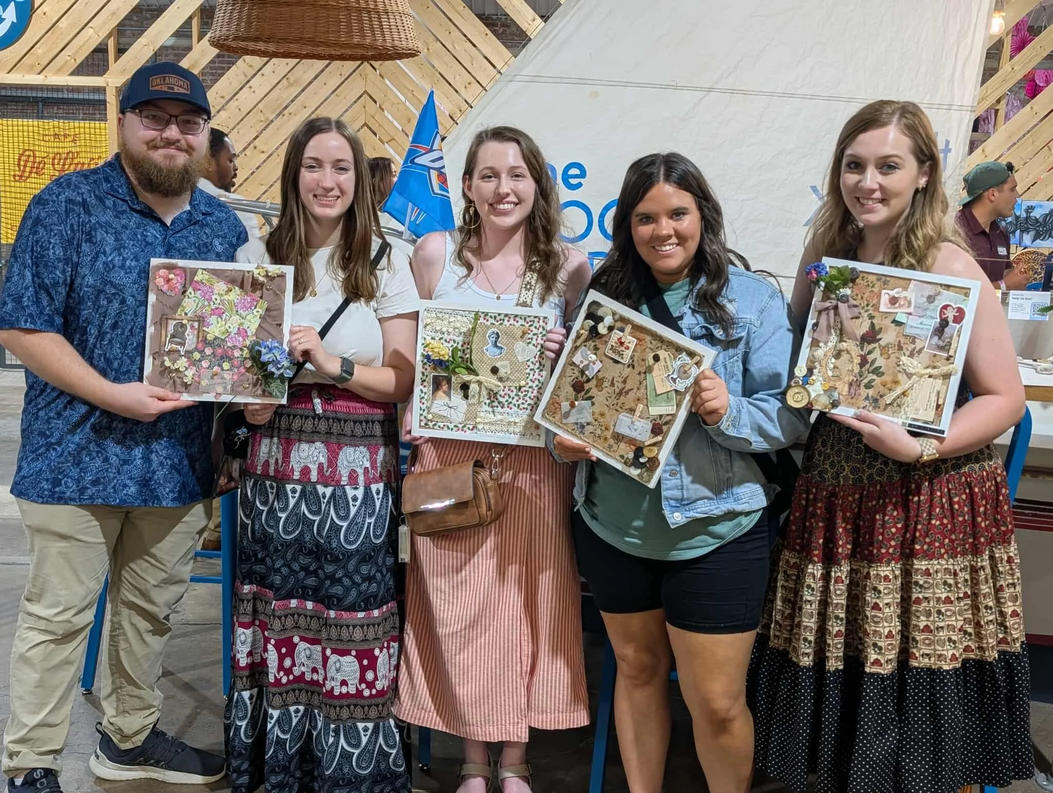 Group of five people standing together holding decorated scrapbook pages at an indoor event.