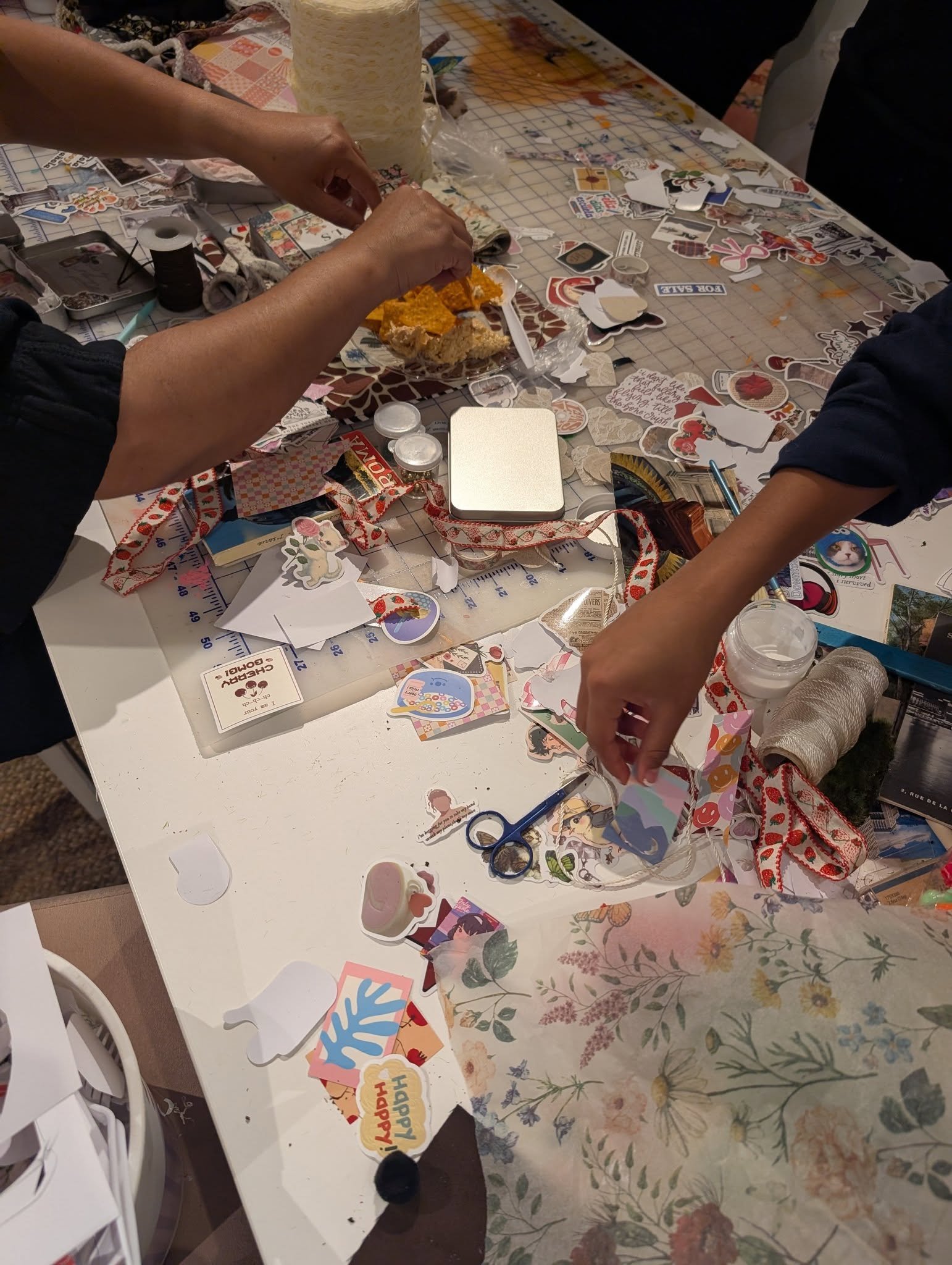 People crafting with stickers, scissors, and decorative paper on a cluttered table for a scrapbooking or card-making activity.
