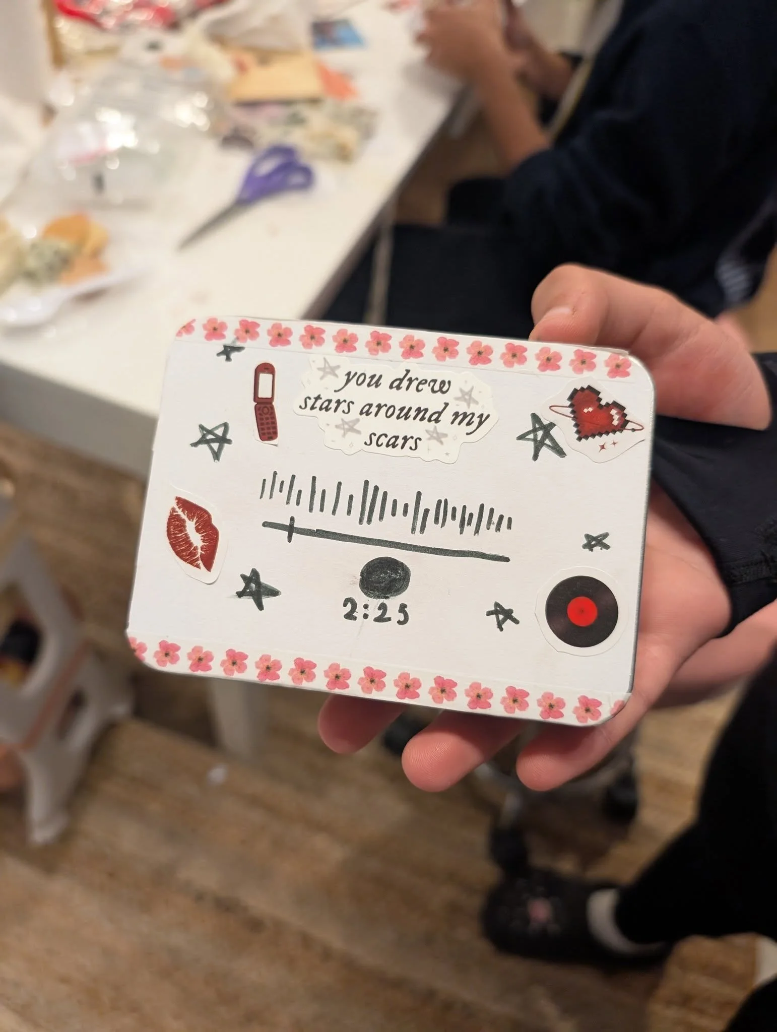 Hand holding a decorated card with drawings and text, including a skateboard, stars, lipstick, a lip print, a heart, a vinyl record, and the message 'you drew stars around my scars' with a timer reading 2:25.