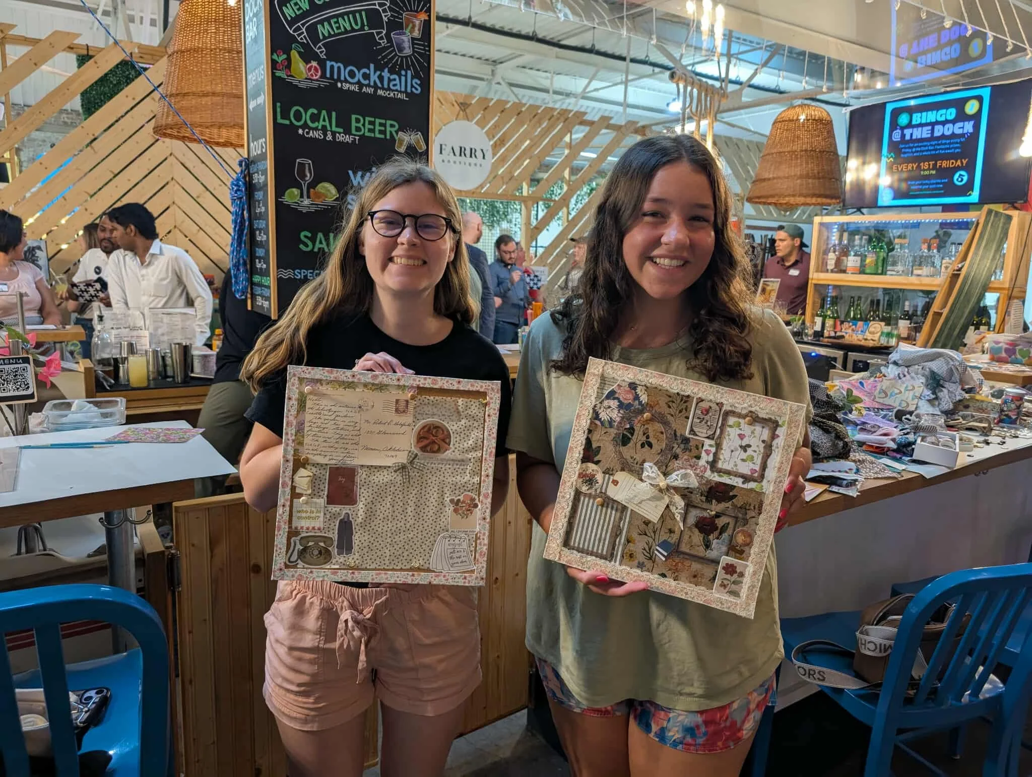 Two young women smiling and holding decorated photo boards inside a lively indoor market or event space with people in the background, a chalkboard menu, and a bar area.