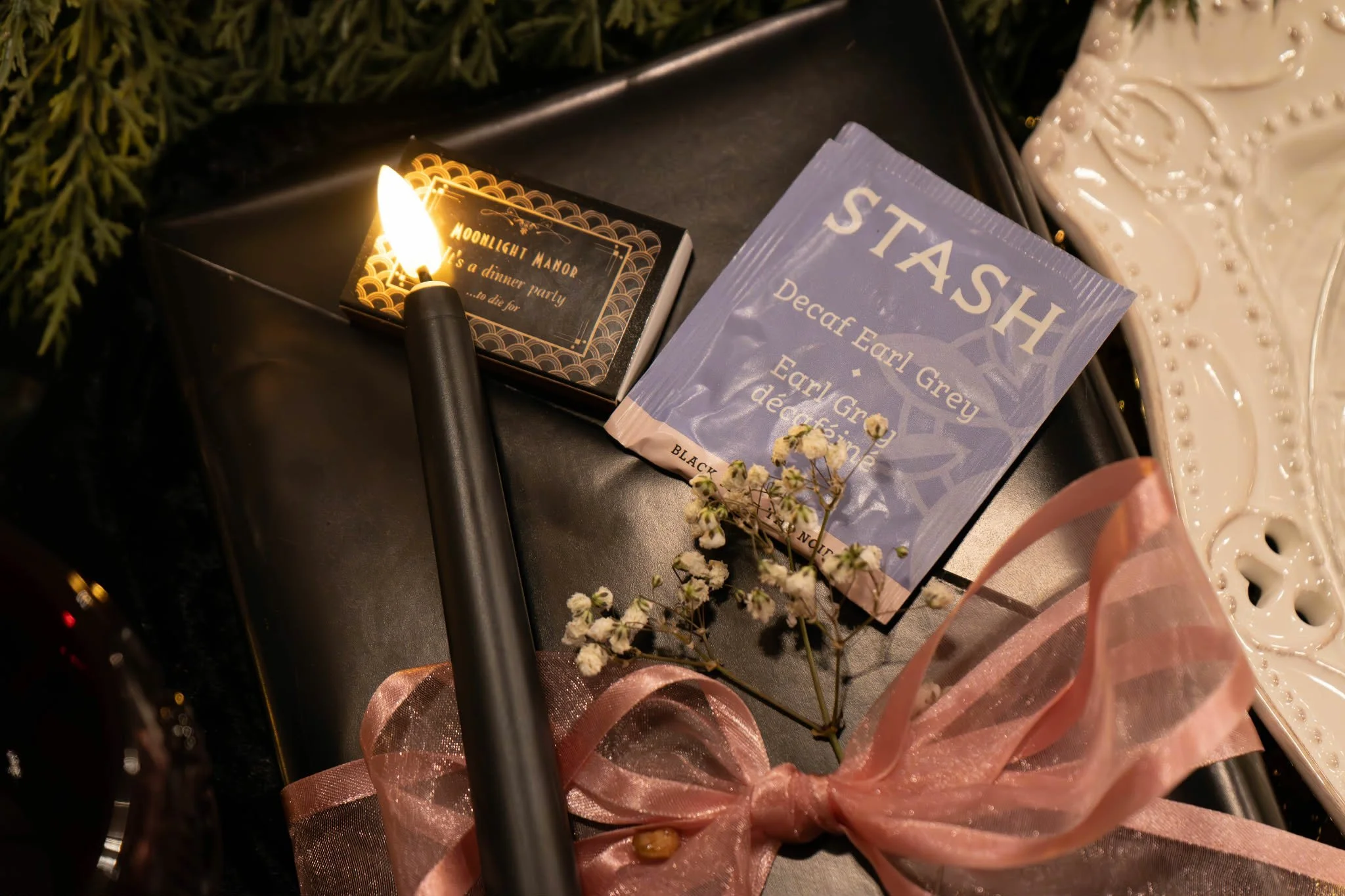 A lit candle, a small box with gold and black design, a packet of black STASH decaf Earl Grey tea, small white flowers tied with pink ribbon, and decorative white ceramic tray on a black surface surrounded by greenery.