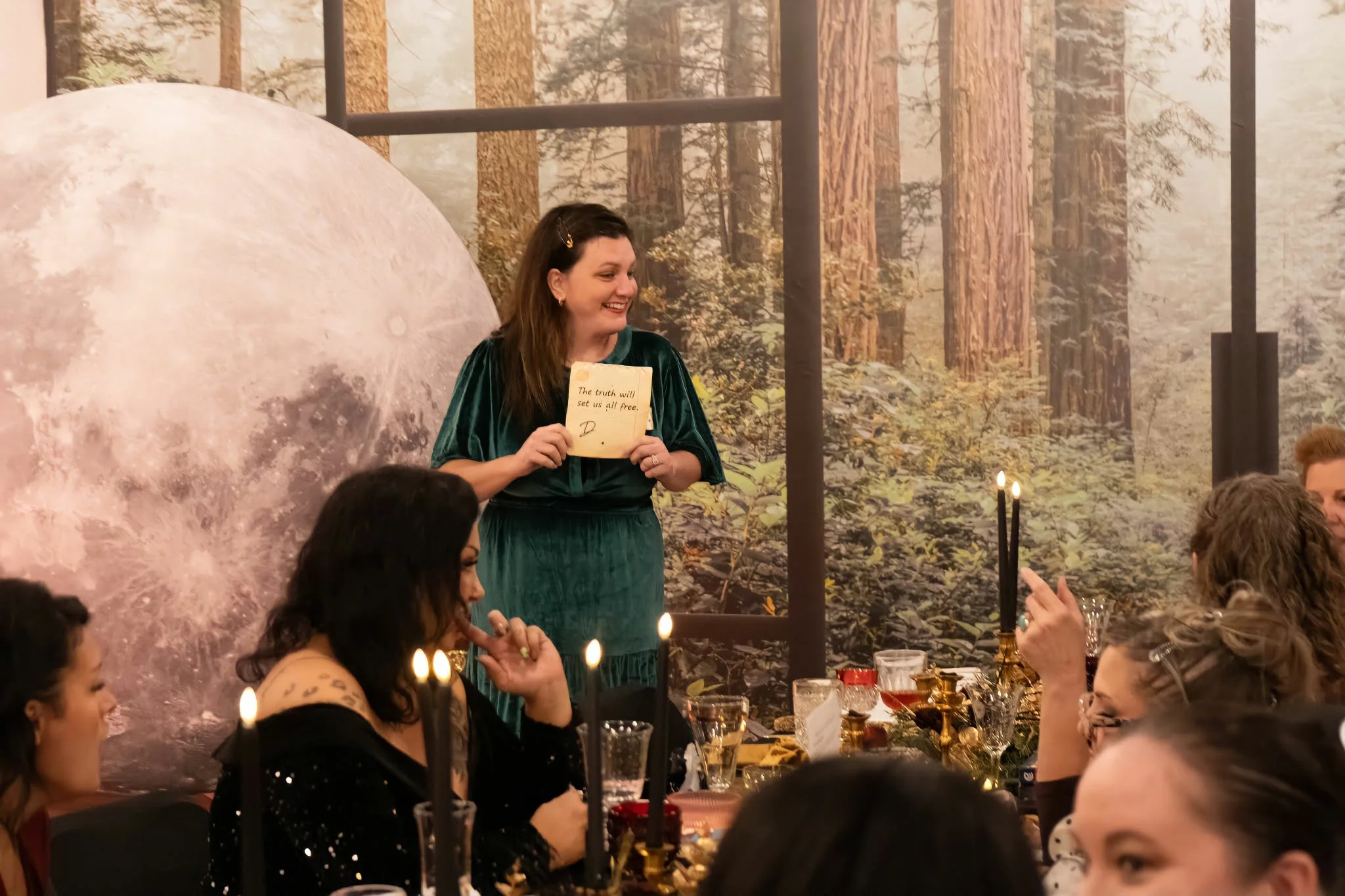 A woman in a green velvet dress standing and smiling at a dinner party, holding a note that says, 'The truth will set us all free.' The background features a forest scene mural and a large image of the moon. Guests are sitting at a table with black candles and ornate tableware.