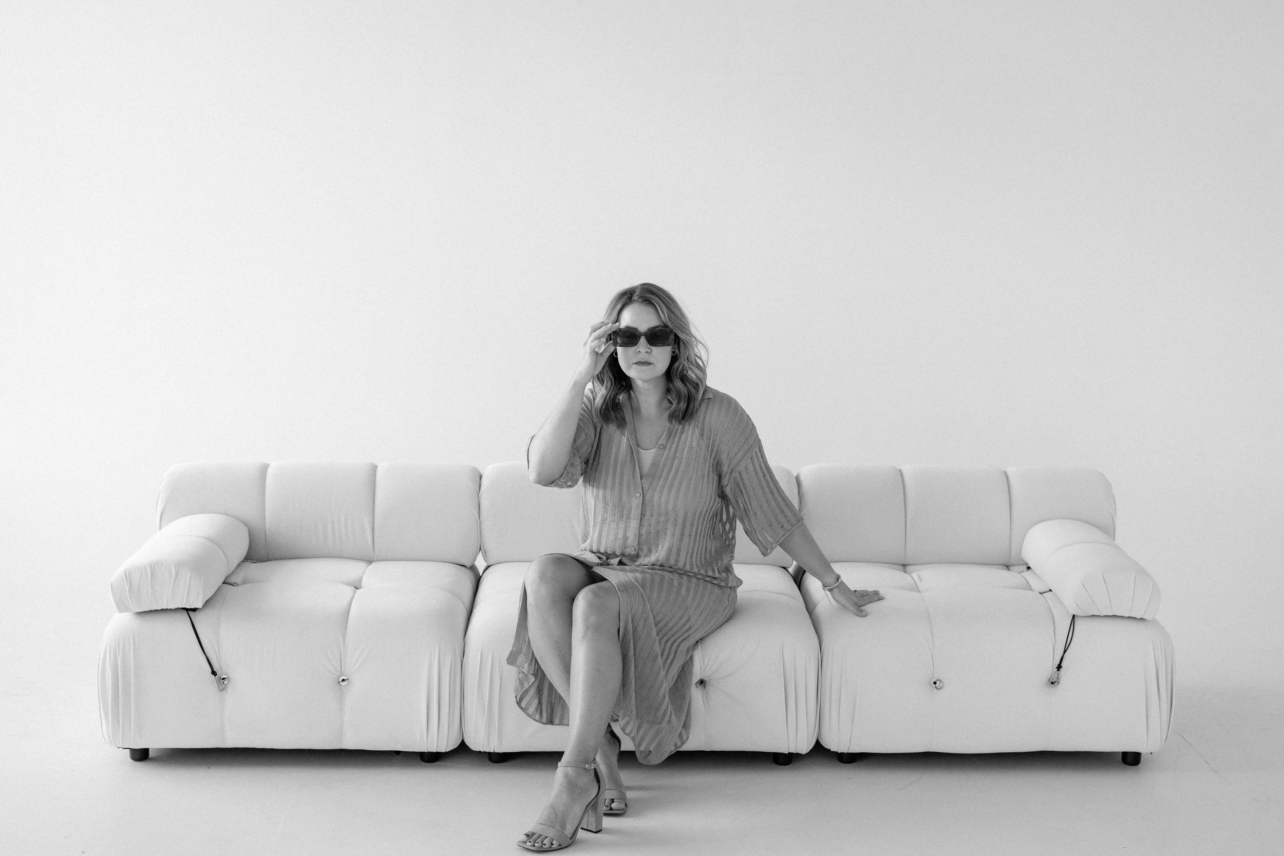 A woman wearing sunglasses and a dress sits on a white leather sofa against a plain white background.