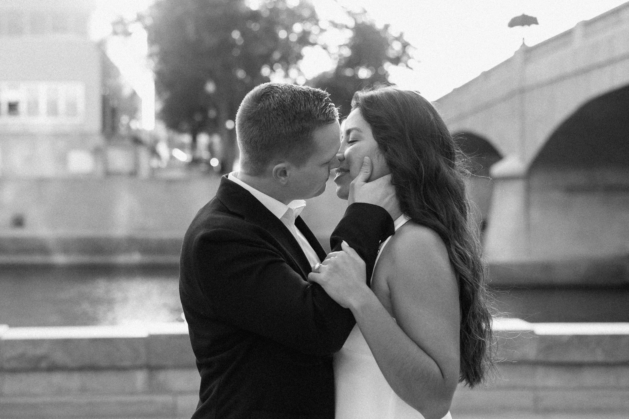 A black and white photo of a couple about to kiss outdoors near a bridge, with trees and buildings in the background.