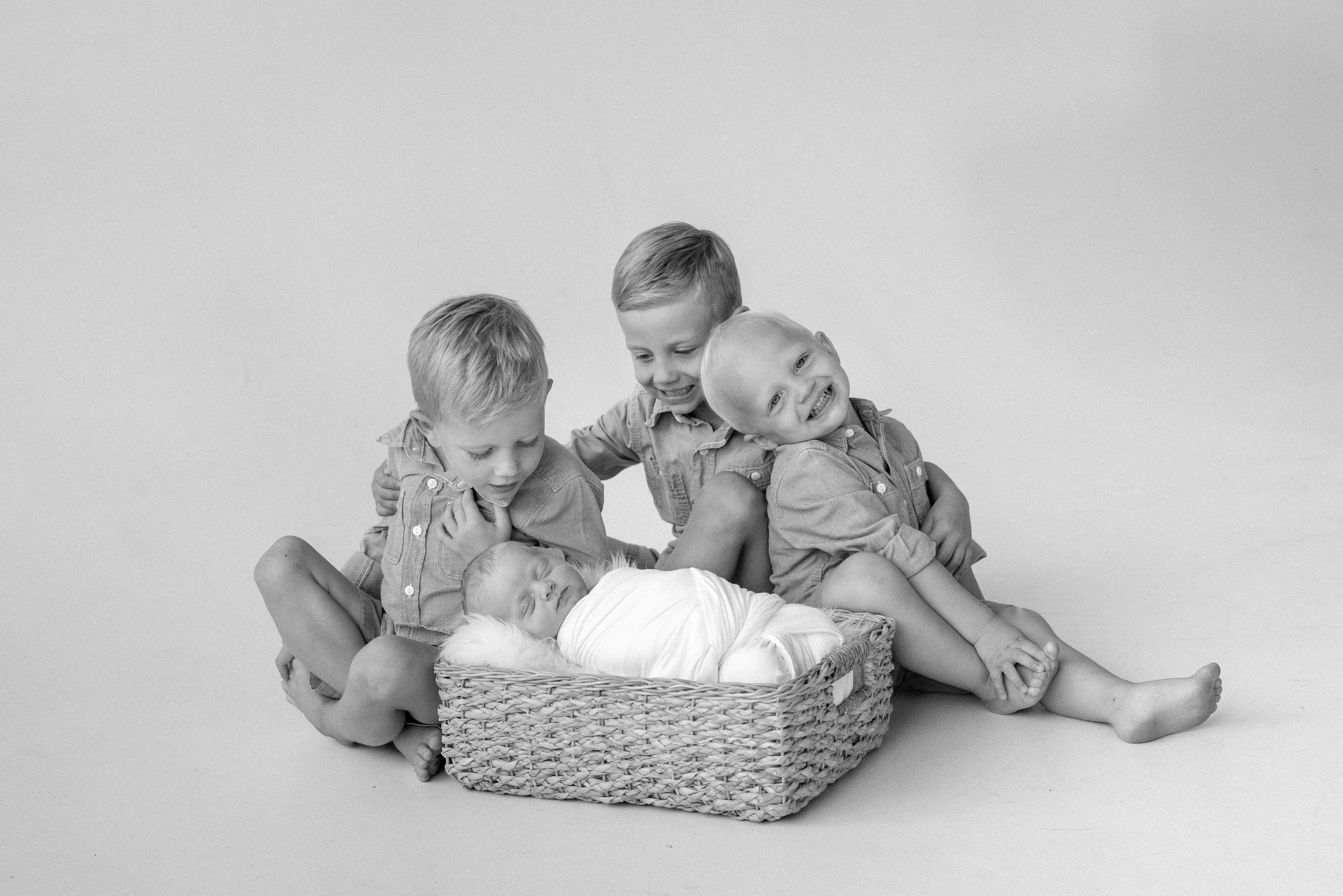 Four young children, three boys and a baby, interact around a sleeping baby in a basket on a plain background.