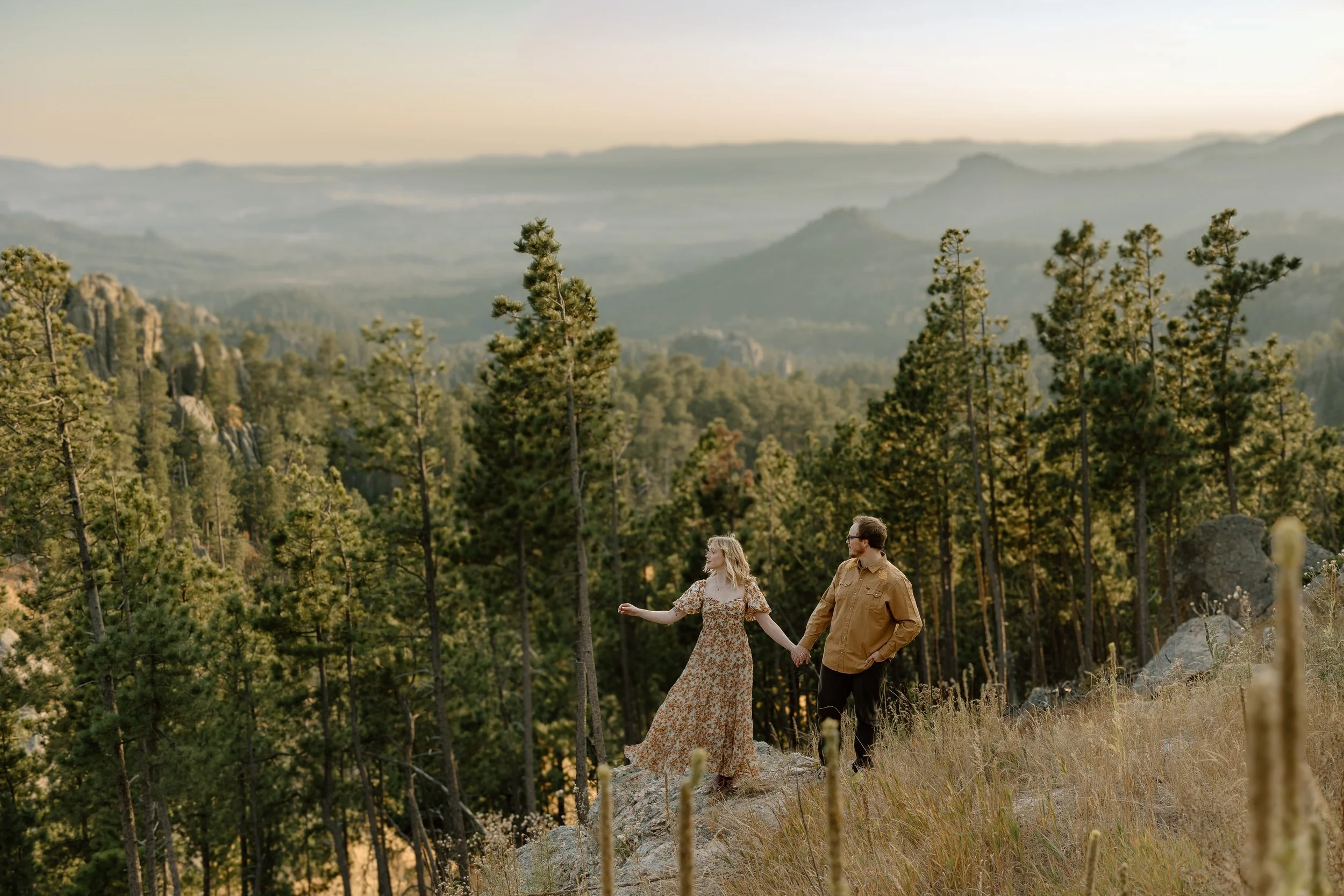 A couple holding hands and walking through a forested landscape during sunset.