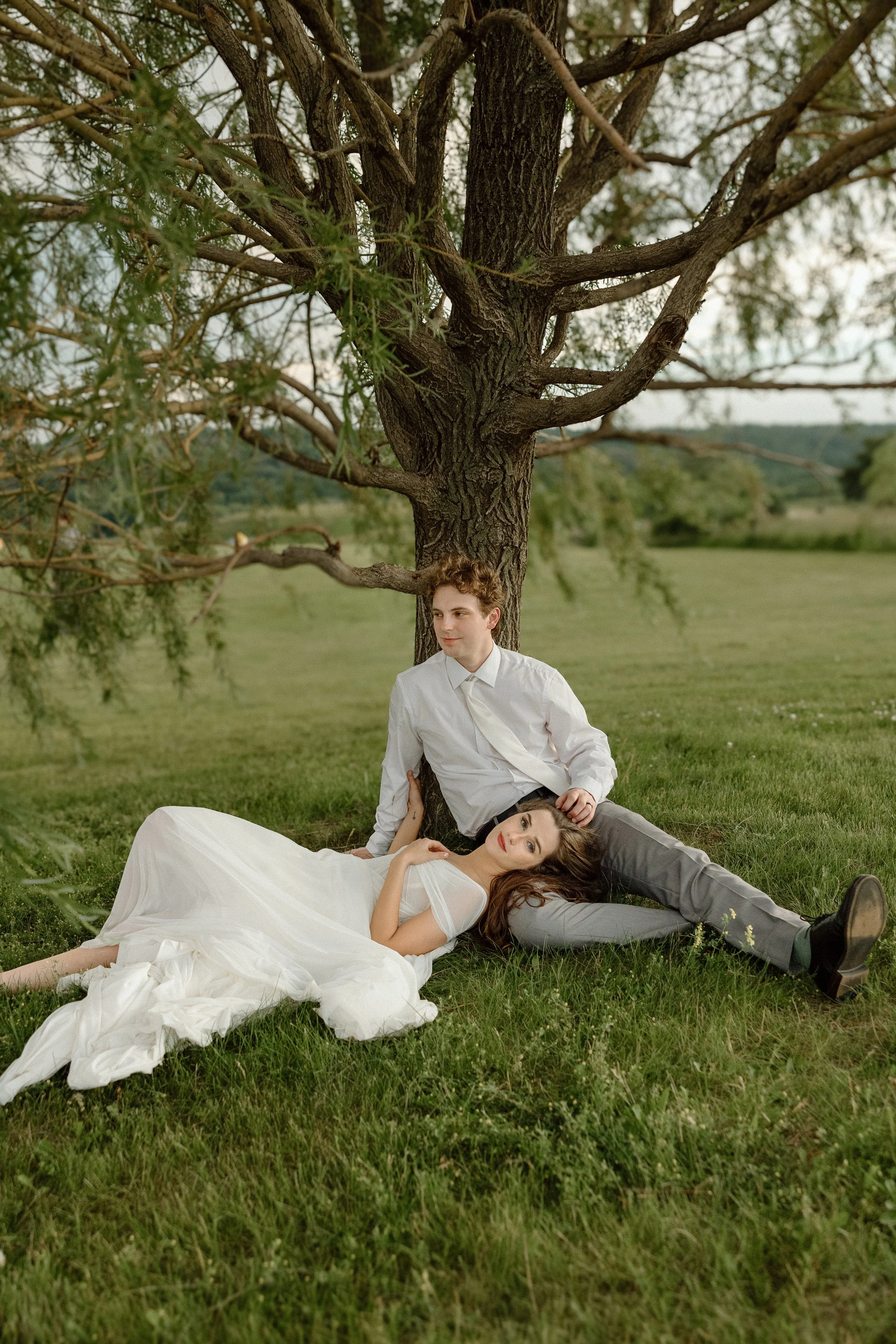 A young man and woman sitting on grass beneath a large tree; the woman is lying down in a white dress, and the man is sitting with his back against the tree, looking to the side.