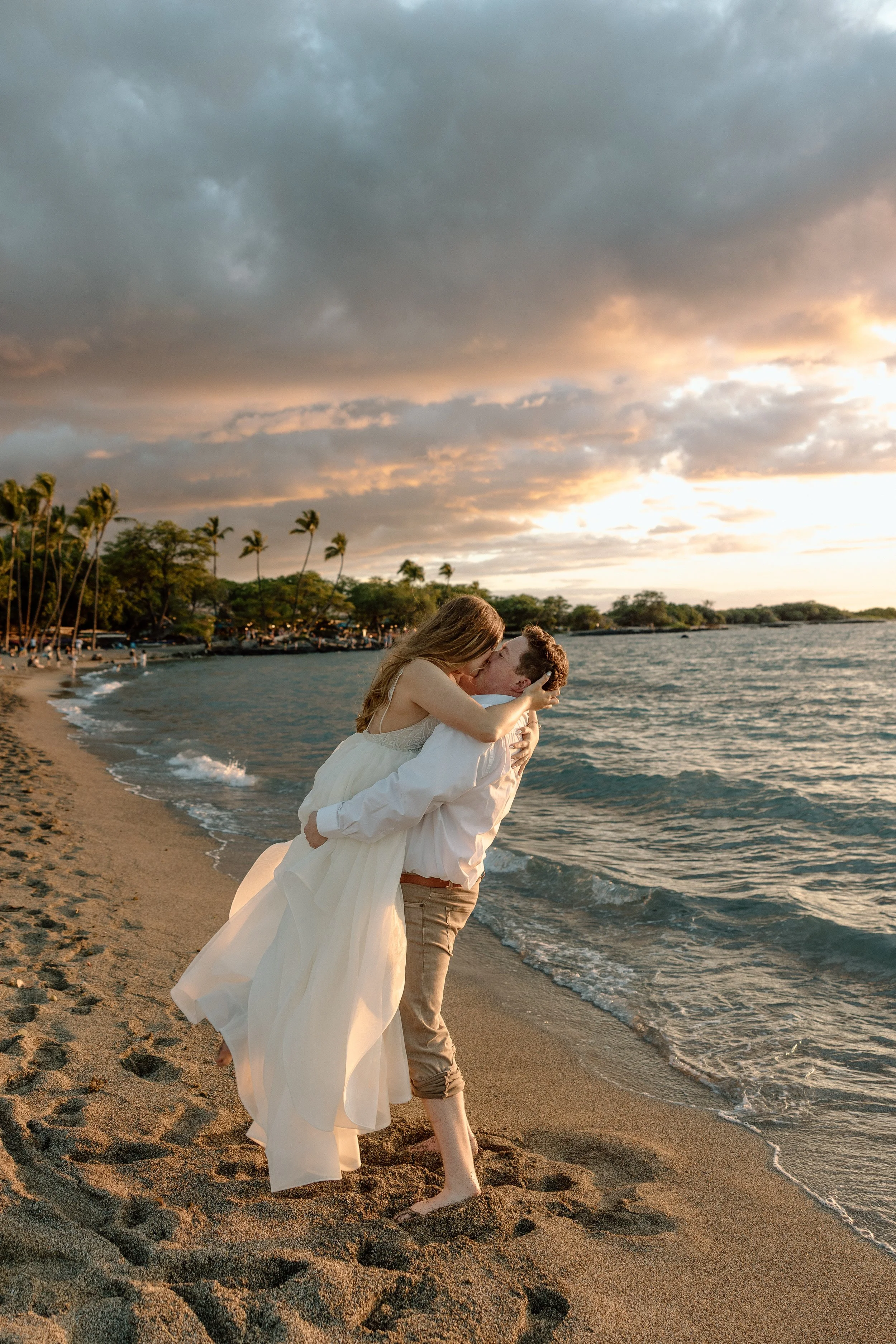 A couple embraces and kisses on a sandy beach at sunset, with palm trees and a cloudy sky in the background.