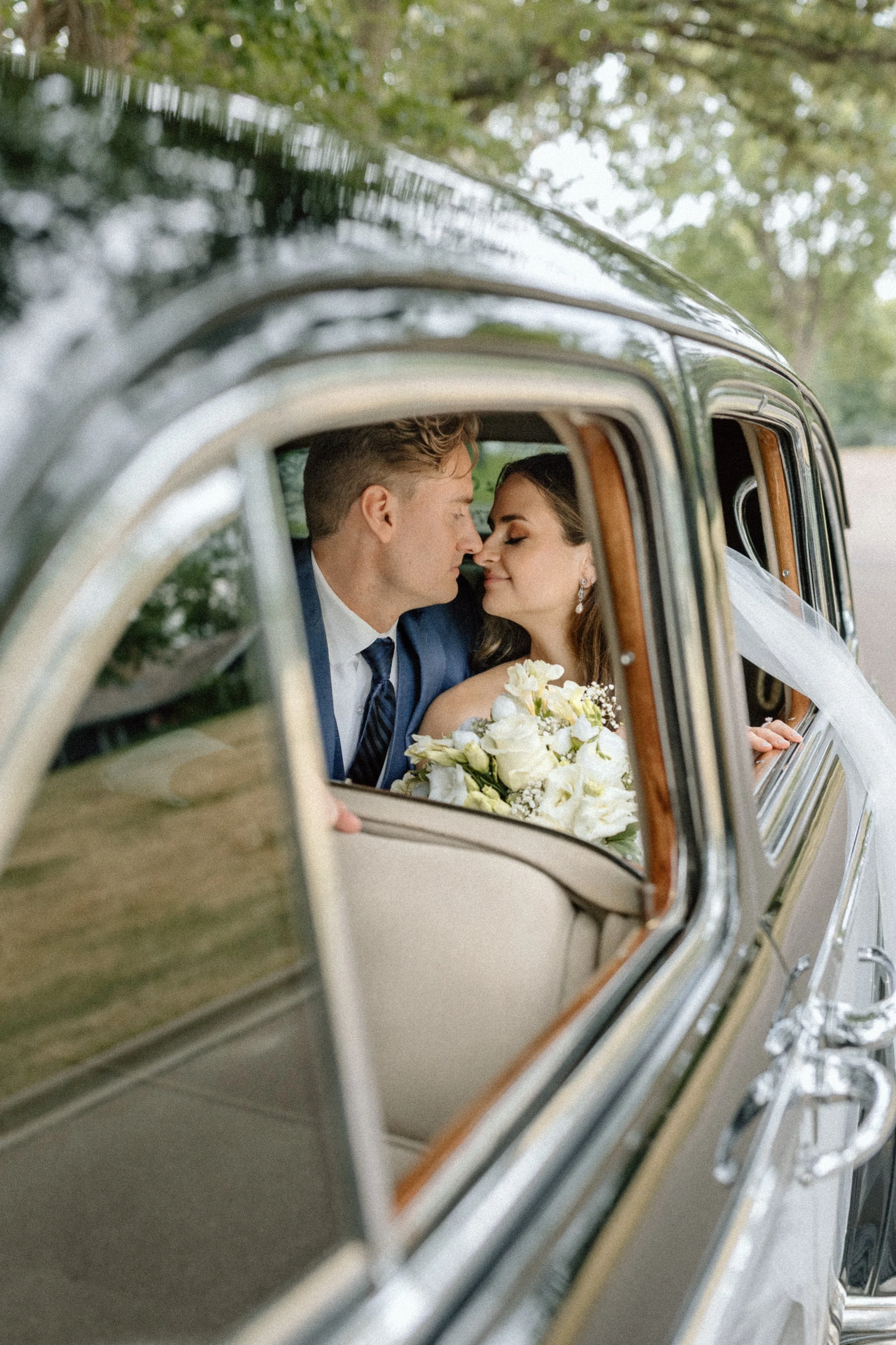 A bride and groom sit close together inside a vintage car, touching noses, with the bride holding a bouquet of white flowers, during a wedding.