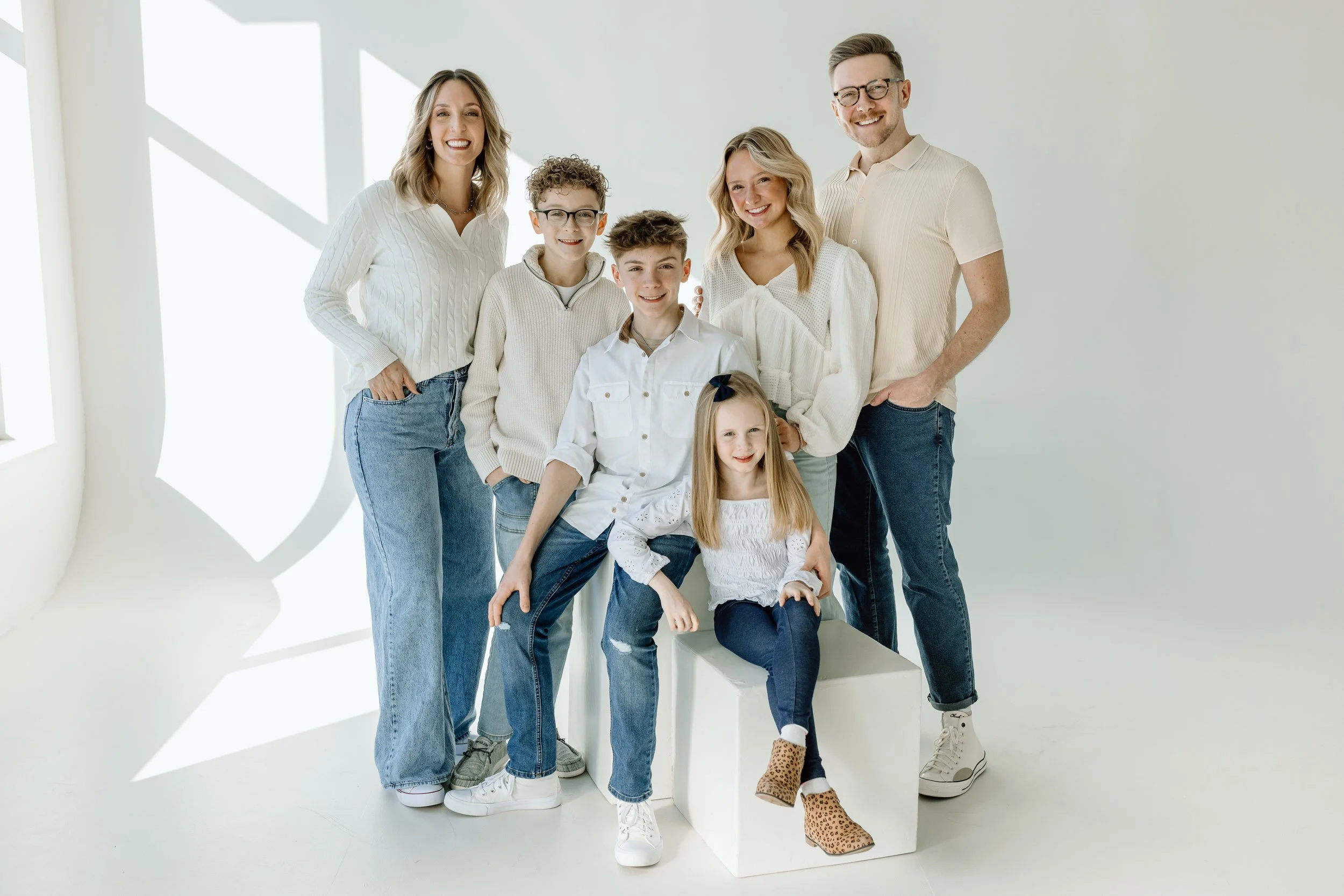 Family portrait of seven people, including three women, two men, and two young girls, standing in a bright, minimalistic white room.