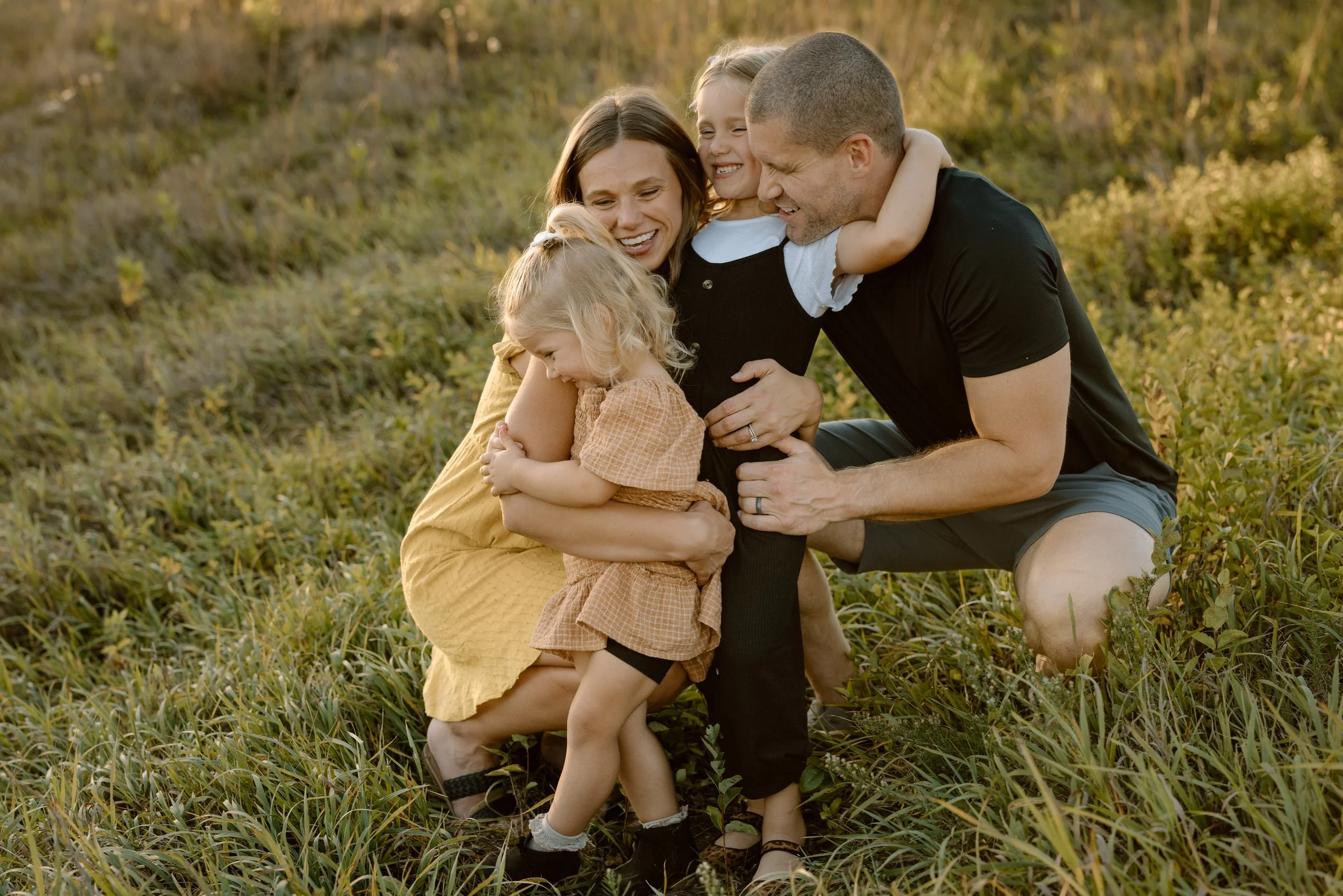 A family of four, two adults and two young girls, hugging and smiling outdoors in a grassy field during sunset.