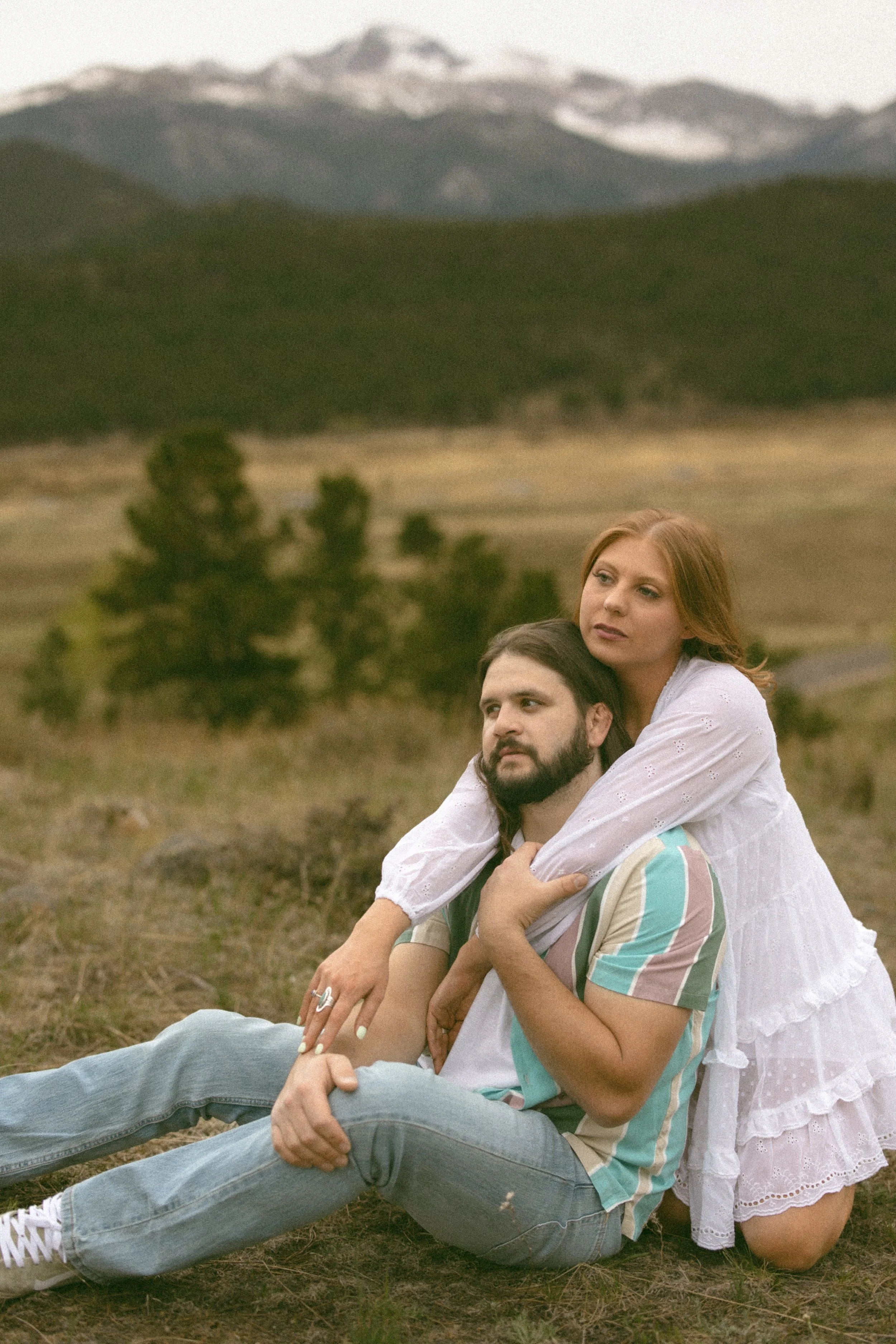 A woman with red hair and a man with dark hair and beard sitting on the ground in a field with mountains in the background. The woman is sitting behind the man, embracing him with her arms around his shoulders, and they both look into the distance.