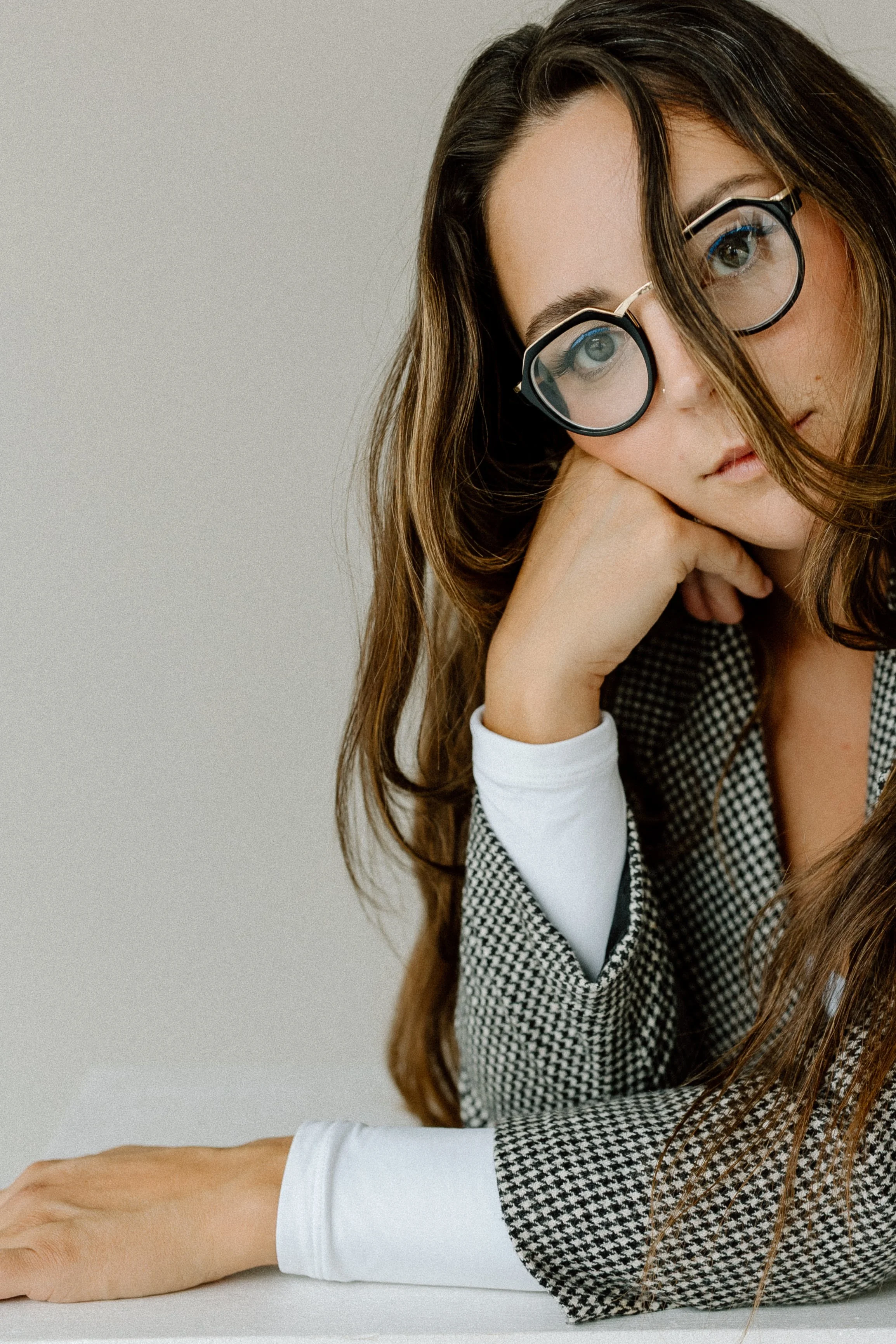 Close-up of a woman with long, wavy brown hair wearing large round glasses, resting her head on her arm, with a calm expression, dressed in a houndstooth blazer and a white shirt, sitting at a table with a plain background.
