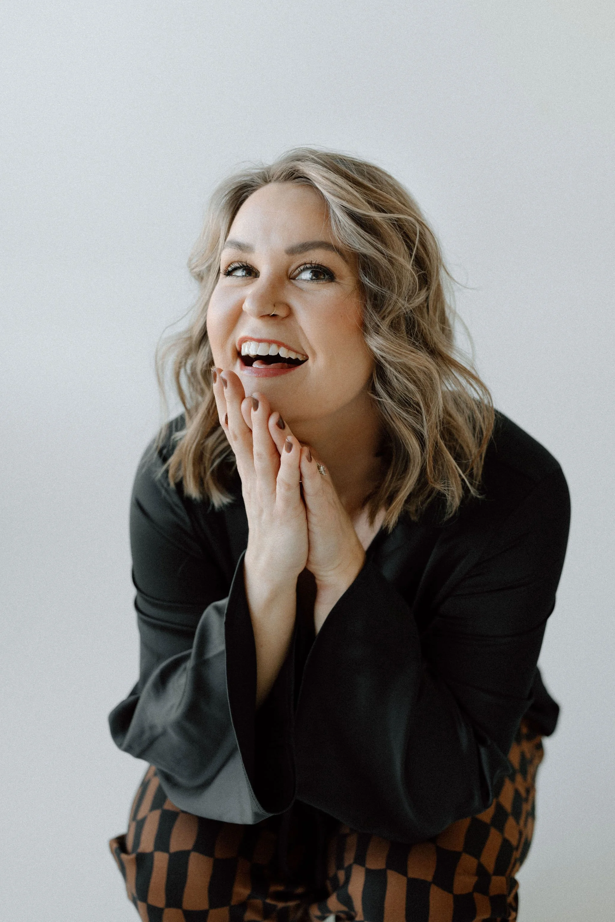 A woman with shoulder-length blonde hair, wearing a black top, is smiling with her hands near her face, looking joyful.