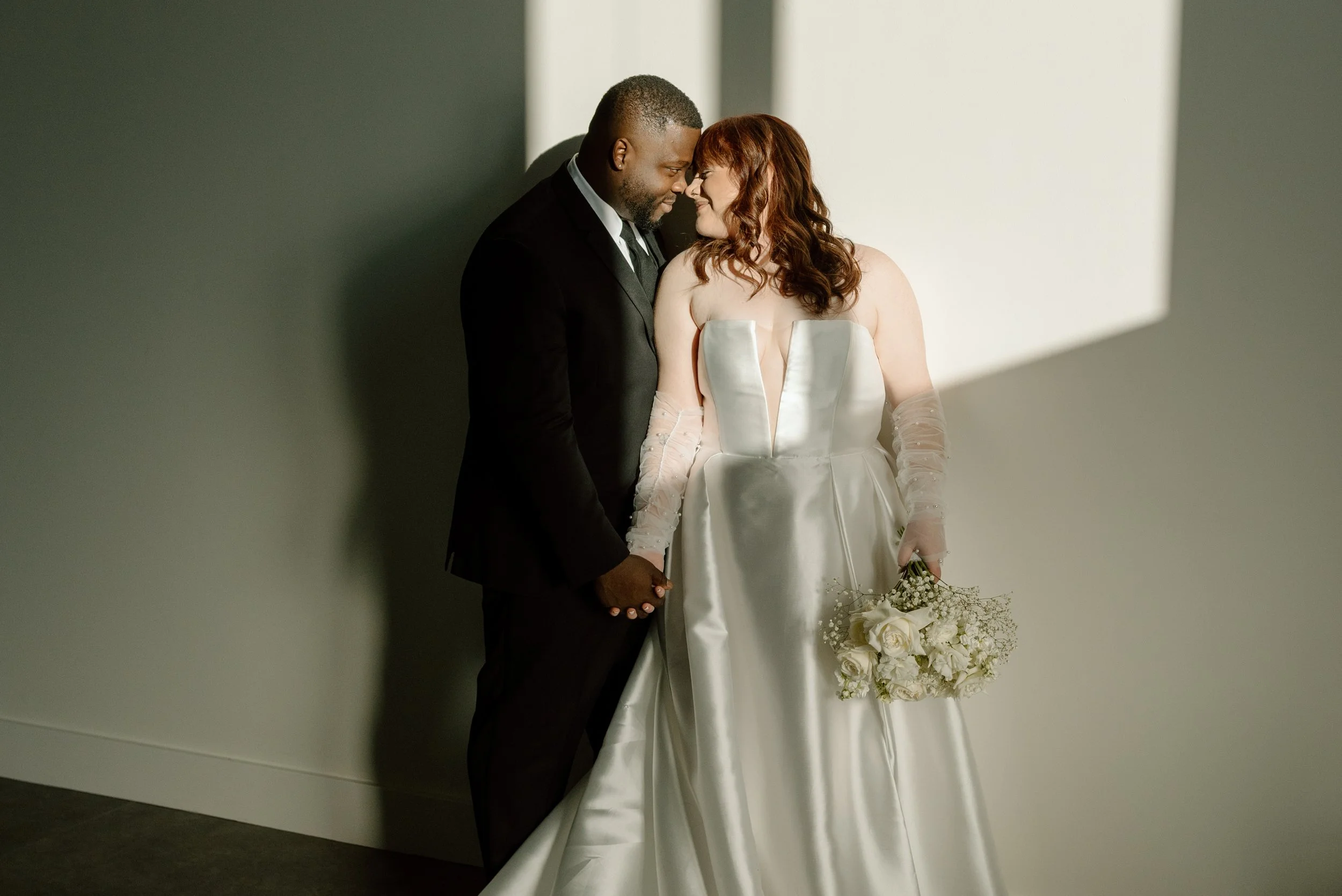 A bride and groom standing close together, holding hands, with their foreheads touching, smiling at each other indoors.