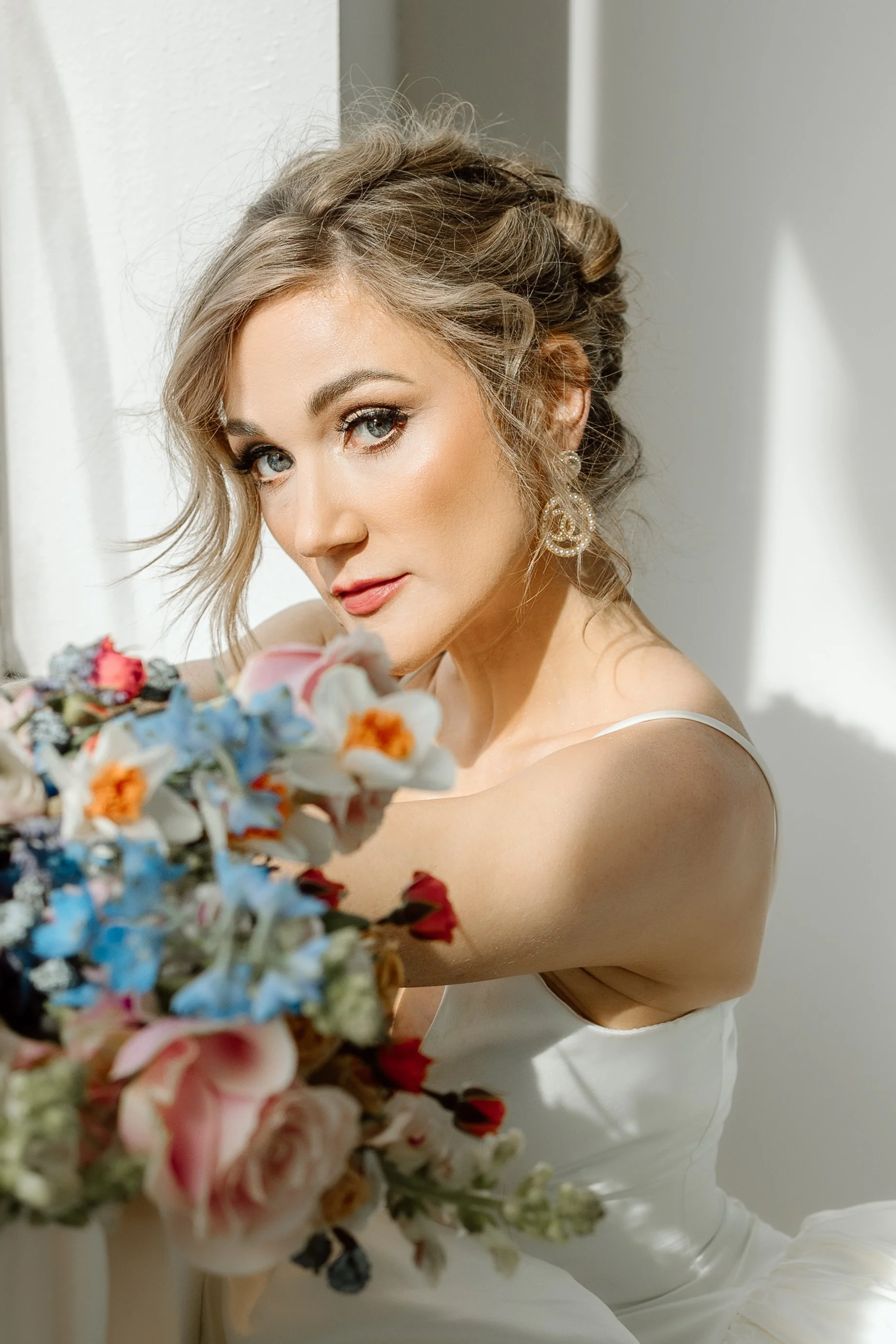 A woman with styled hair, makeup, and pearl earrings looking at the camera while holding a bouquet of colorful flowers.