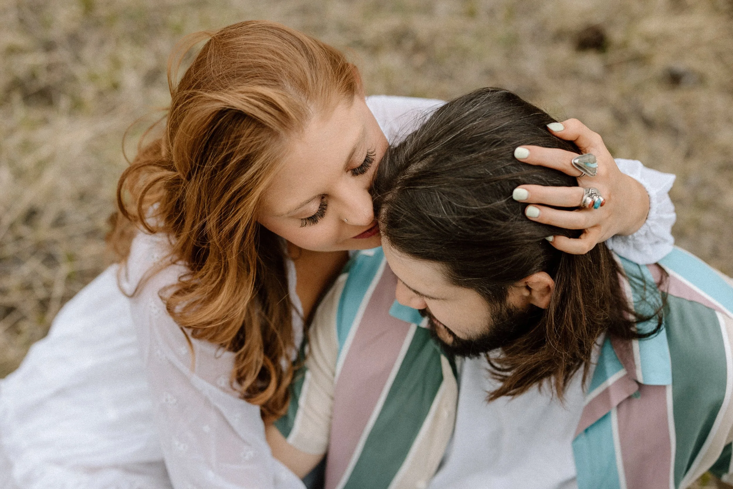 A woman with red hair gently holds and kisses a man with dark hair and a beard on his forehead, both with their eyes closed, outdoors on a grassy surface.