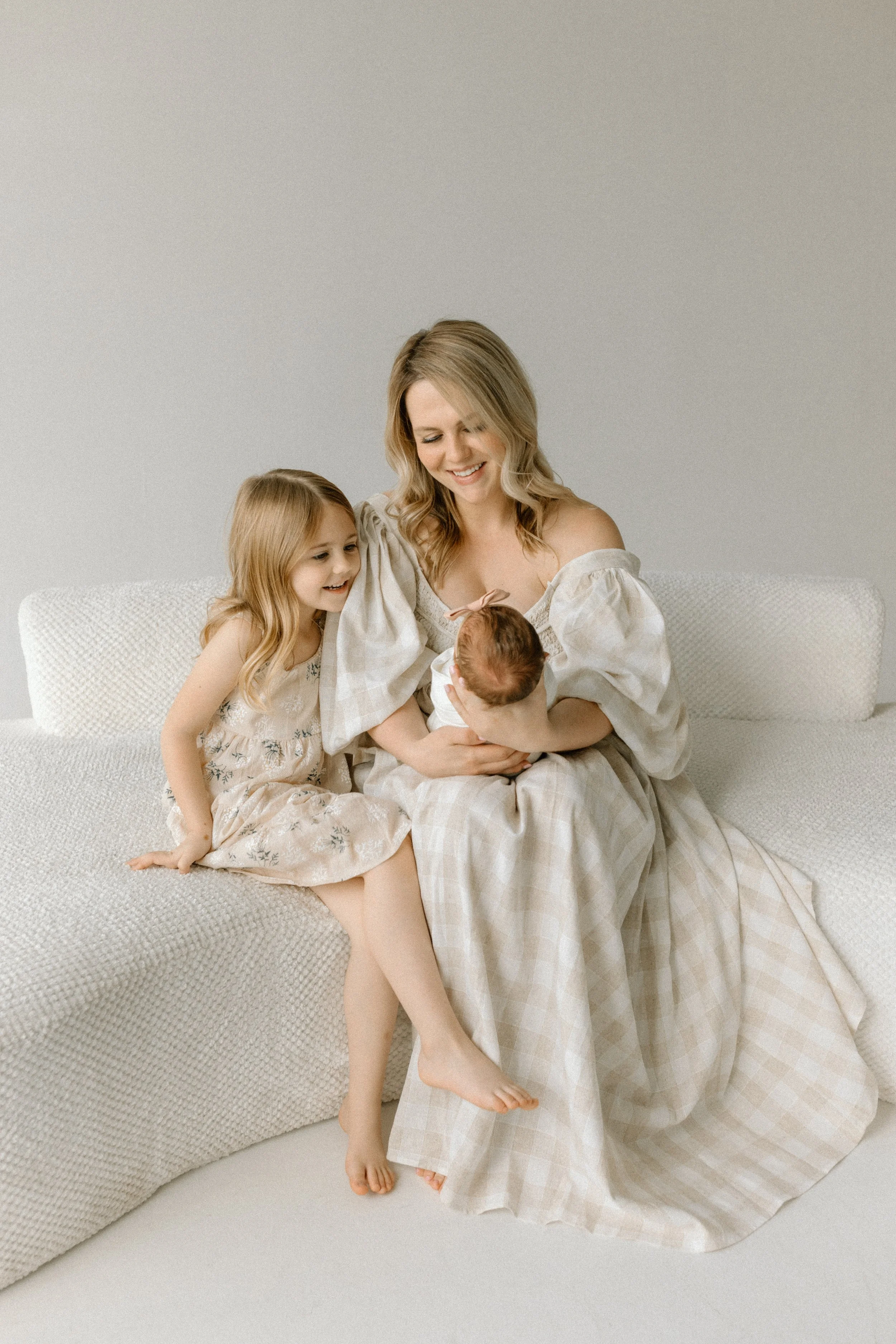 A woman with blonde hair and two young girls sitting on a white textured sofa in a neutral-colored room. The woman is holding a newborn baby while the older girl sits close, all three looking at the baby with smiles.
