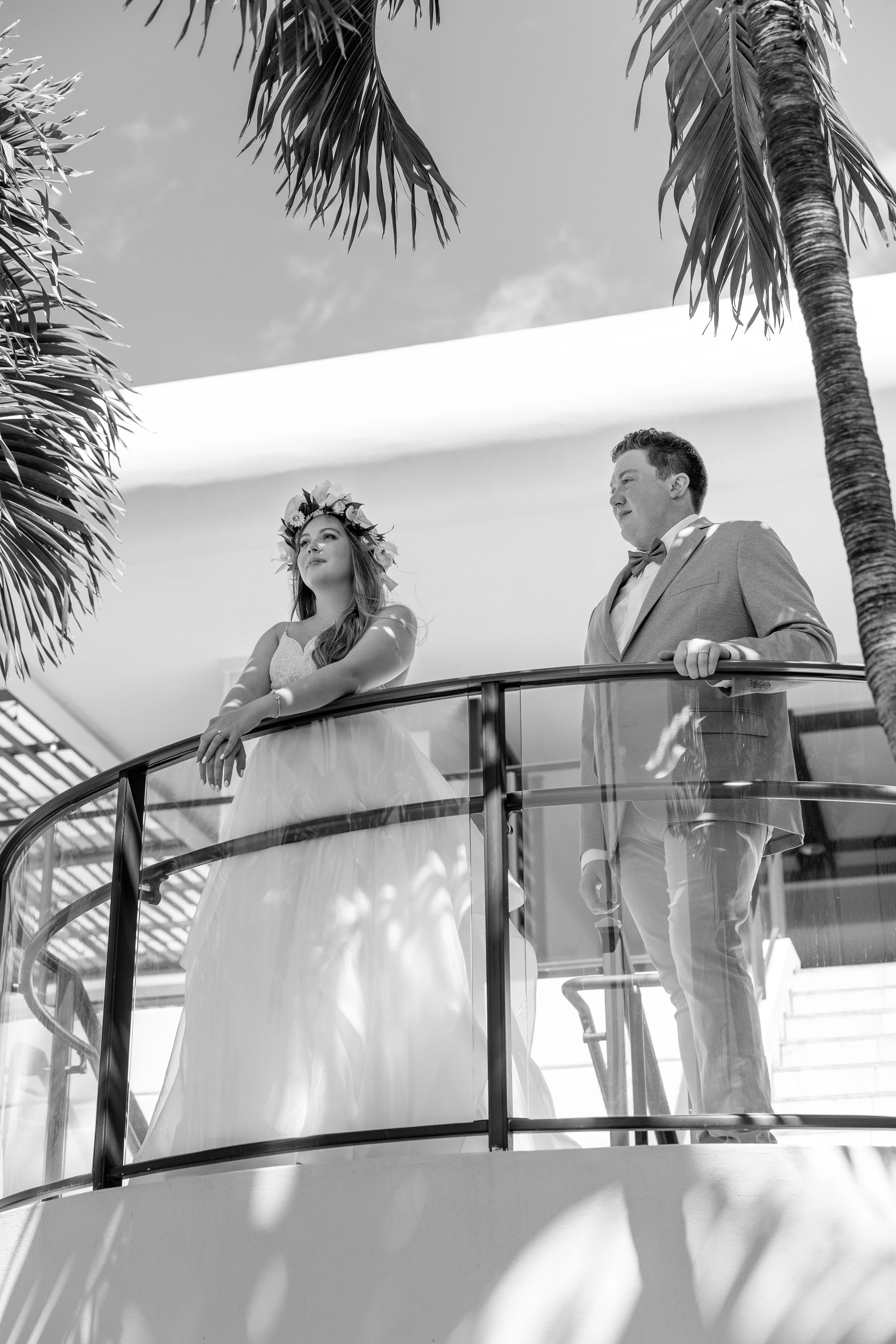 Bride and groom standing on a balcony during a wedding ceremony, with palm trees and a clear sky in the background, in black and white.