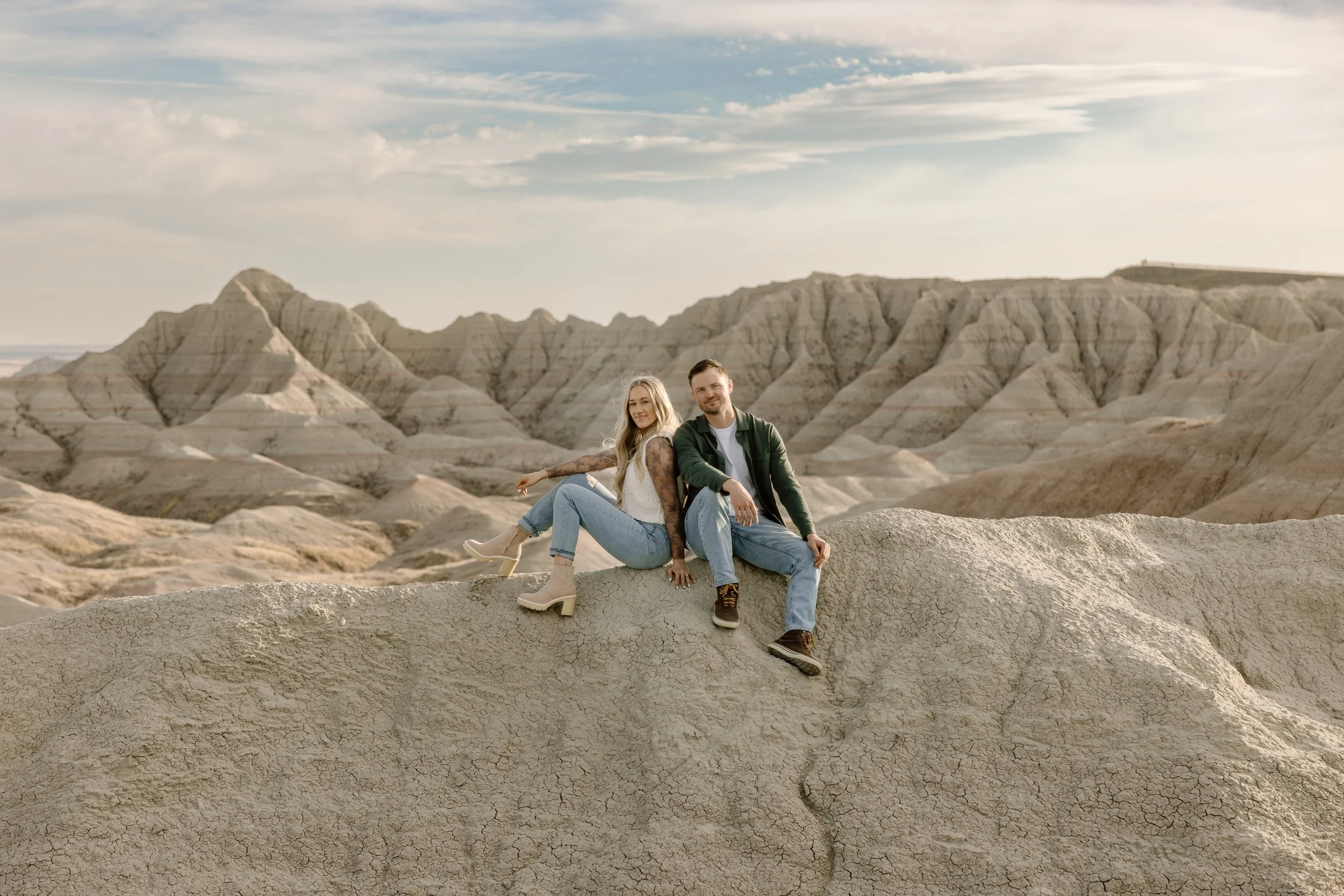 A man and woman sitting on a large rock formation with petrified, layered mountains in the background under a partly cloudy sky.